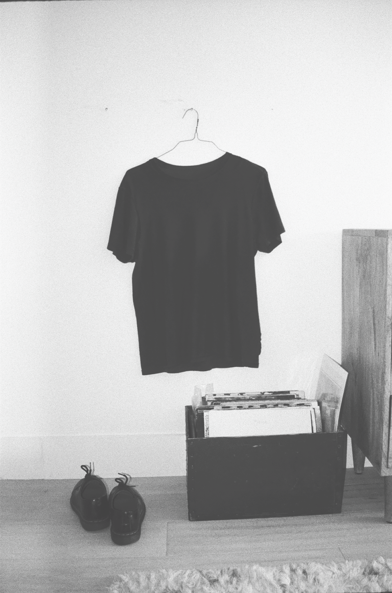 Black T-shirt hanging on a wire hanger on a plain white wall. Below, a pair of black shoes on the floor, and a wooden storage box filled with records.