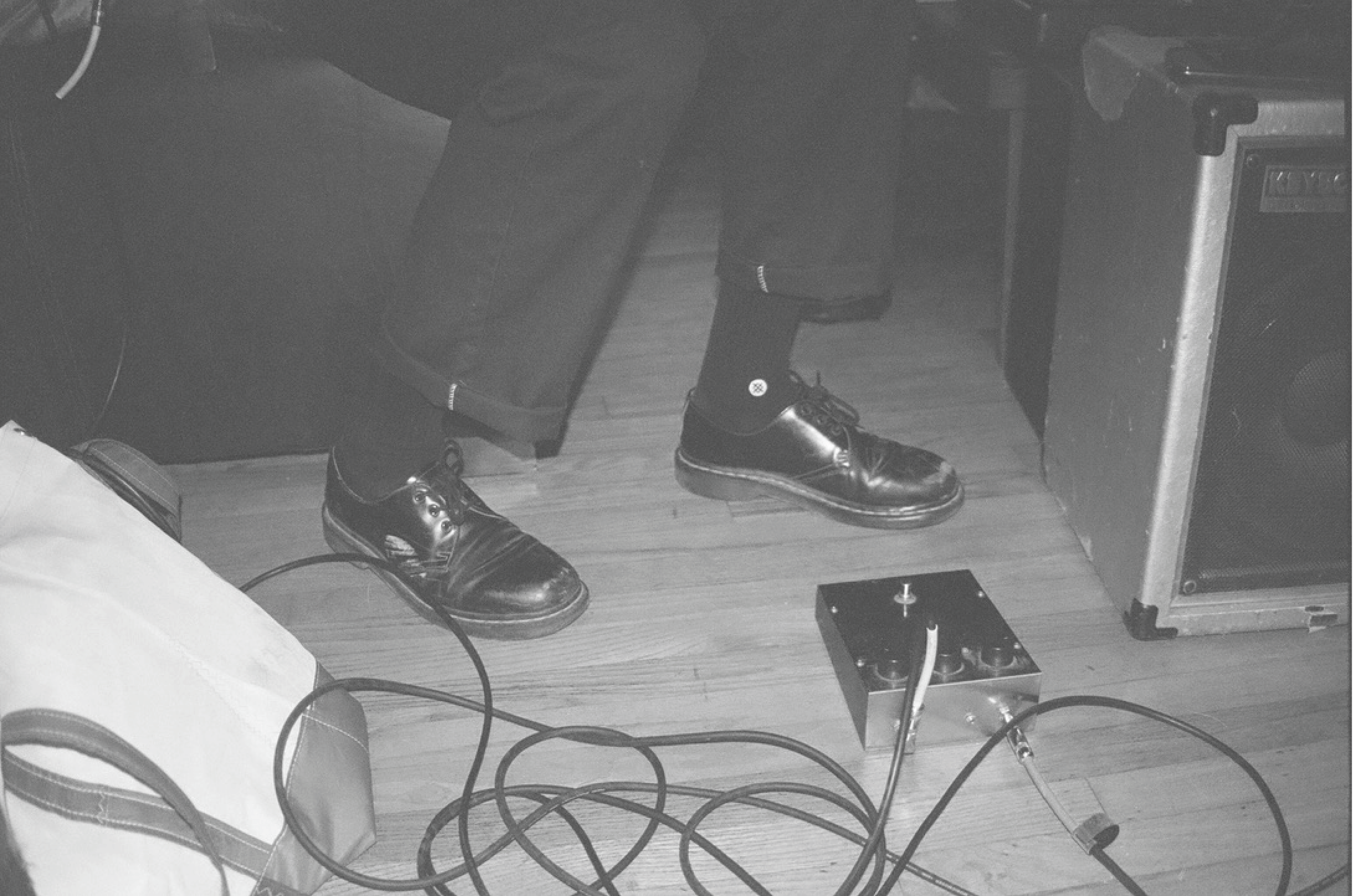 Close-up of a person's black leather shoes, black socks, and black pants, standing on a wood floor. Various electronic equipment and cables are scattered around, including a metal box with a cable attached, and large speakers.