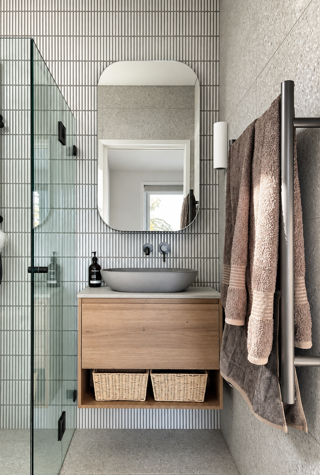 Modern bathroom with a floating wooden vanity, oval sink, wall-mounted faucet, large mirror, brown towels on a towel rack, and a glass shower door. Neutral tones and textured wall tiles.