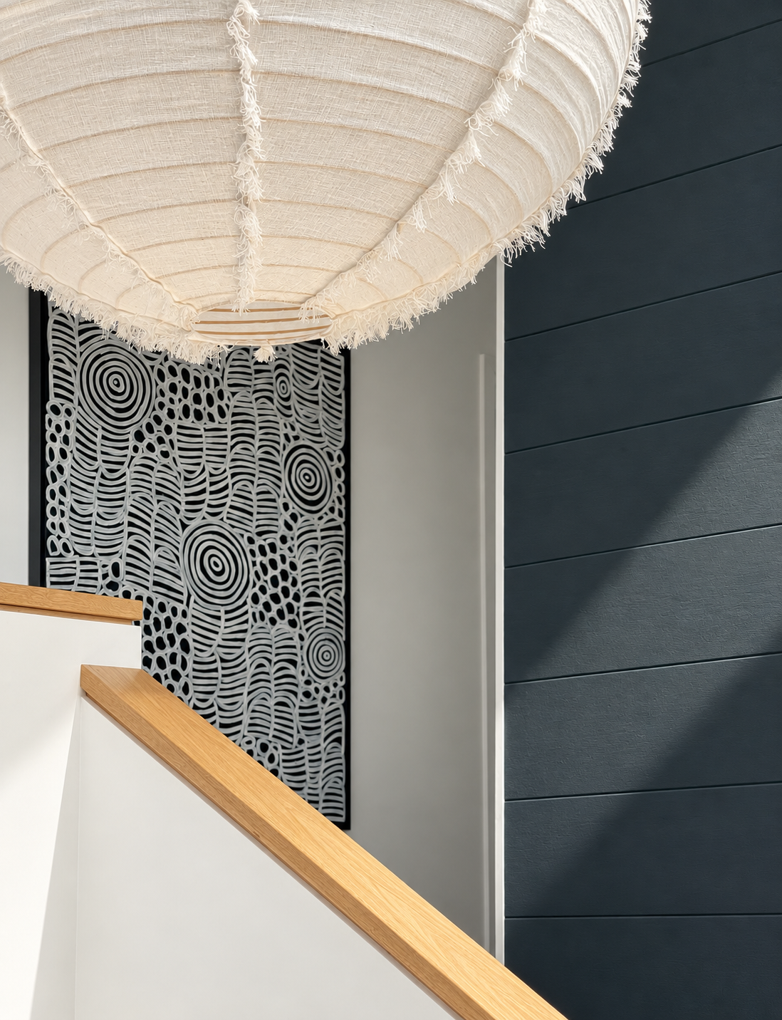 Close-up of an indoor staircase with wood handrail, black patterned wall art, and dark gray horizontal wall panels, illuminated by a large beige fabric lampshade.