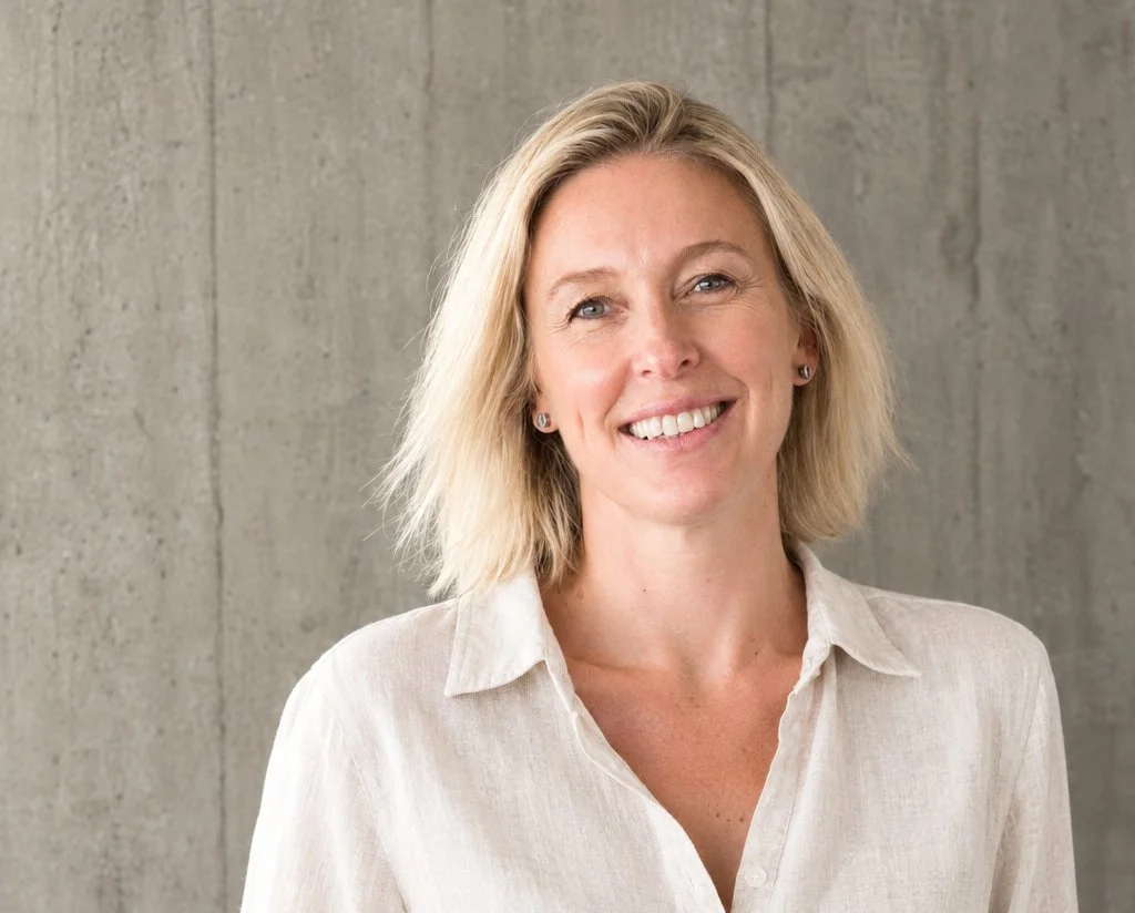 Portrait of a smiling woman with shoulder-length blond hair, wearing a cream-colored blouse, standing against a gray concrete wall.