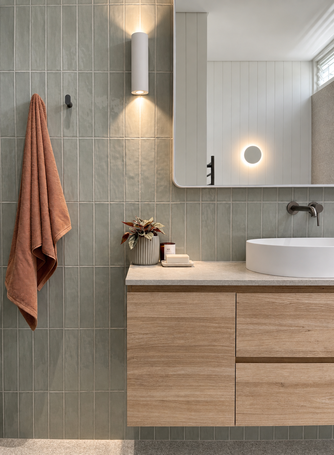 Bathroom with a large mirror, green tiled wall, white vessel sink, wall-mounted faucet, light wood vanity, pink towel hanging on wall hook, decorative plant, candle, soap on countertop, wall-mounted light fixture, and ceiling light.