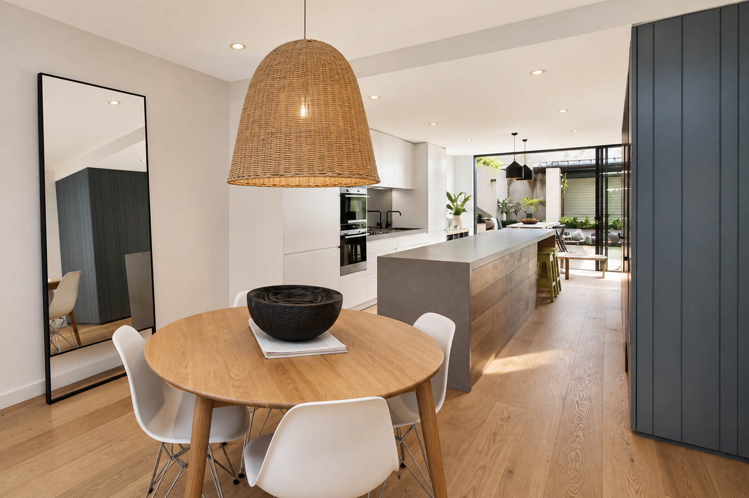 Modern open-concept kitchen and dining area with a round wooden table, white chairs, large hanging wicker light, white cabinetry, and a kitchen island with bar stools, leading to an outdoor patio.