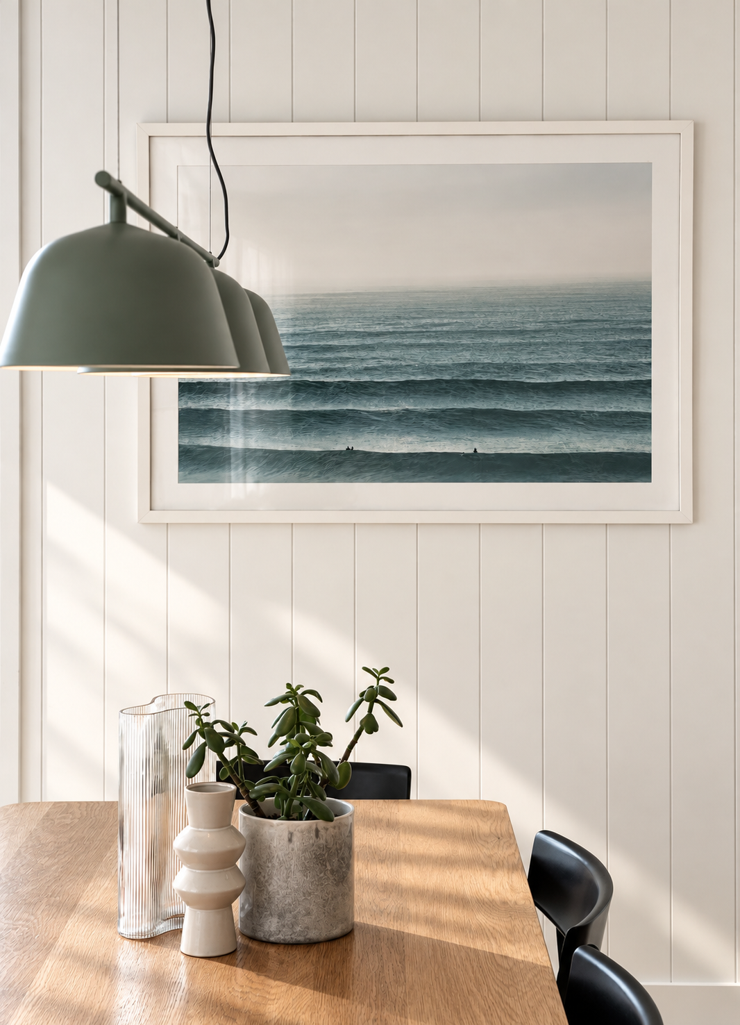 A dining room with a wooden table, black chairs, a large framed ocean photograph on wainscoted wall, two plants and decorative vases on the table, and a green pendant lamp hanging overhead.