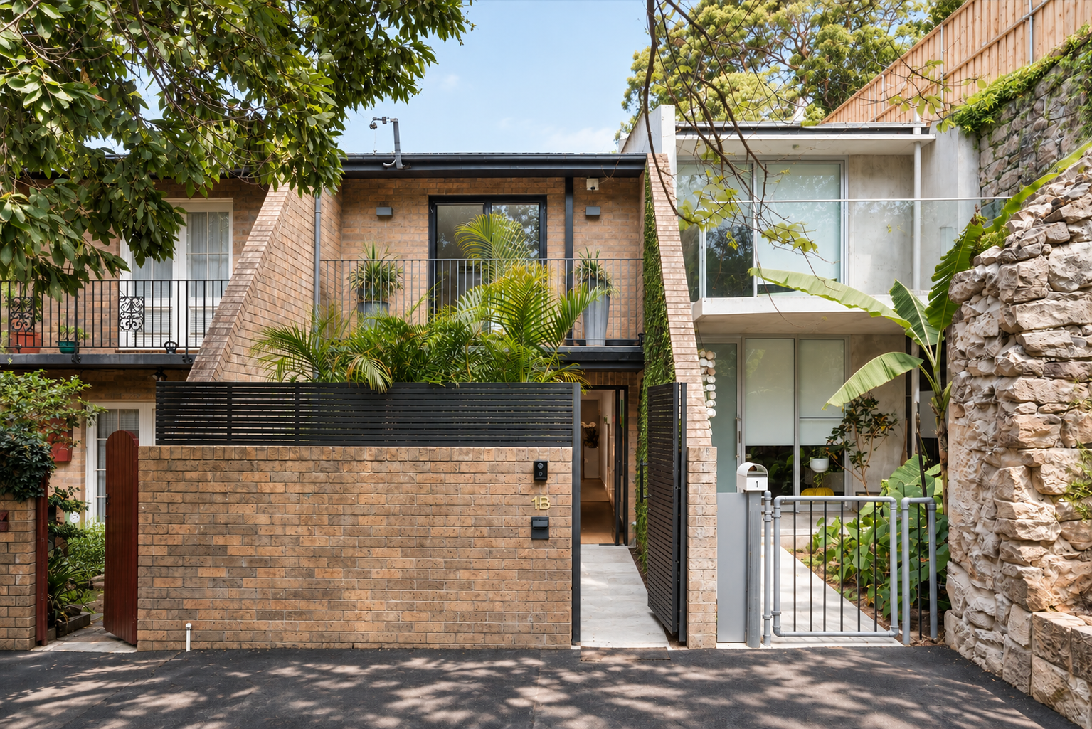 Modern two-story brick residential building with urban garden, balcony, metal gate, and lush greenery.