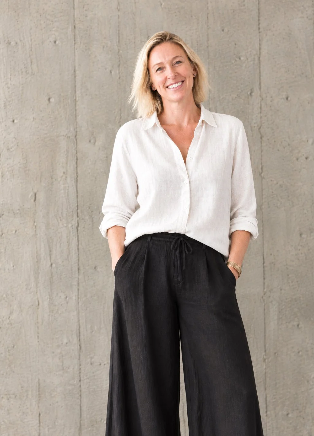 A smiling woman with shoulder-length blonde hair, wearing a white button-up shirt with rolled-up sleeves and black wide-leg pants, stands in front of a concrete wall with her hands in her pockets.