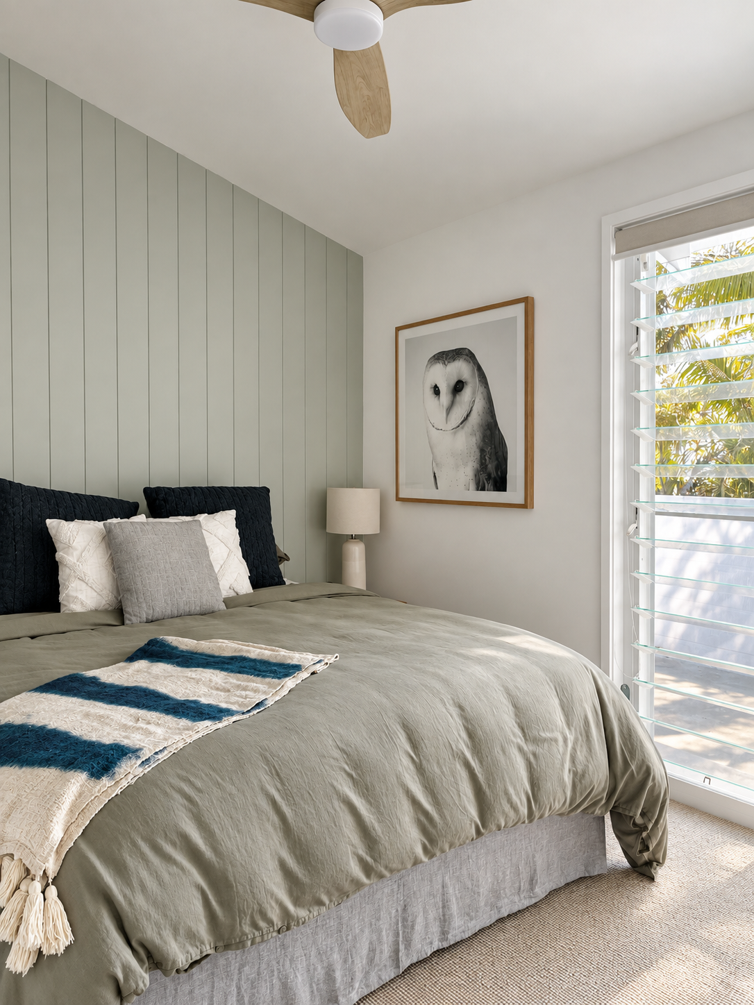 A bedroom with a bed featuring beige bedding, four pillows, and a blue-and-white throw blanket. There is a lamp on the nightstand, a framed owl artwork on the wall, and a window with white shutters letting in sunlight and showing outdoor foliage.