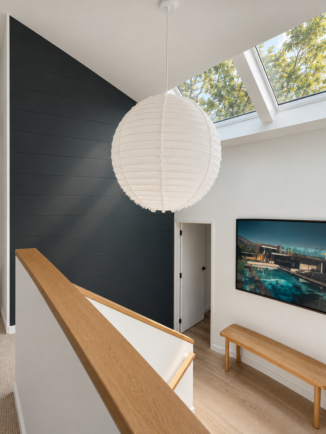 Interior of a modern home with a large white paper lantern hanging from the ceiling, a dark gray accent wall, skylight with a view of green trees, a wooden bench, and a framed picture of a house with a pool.
