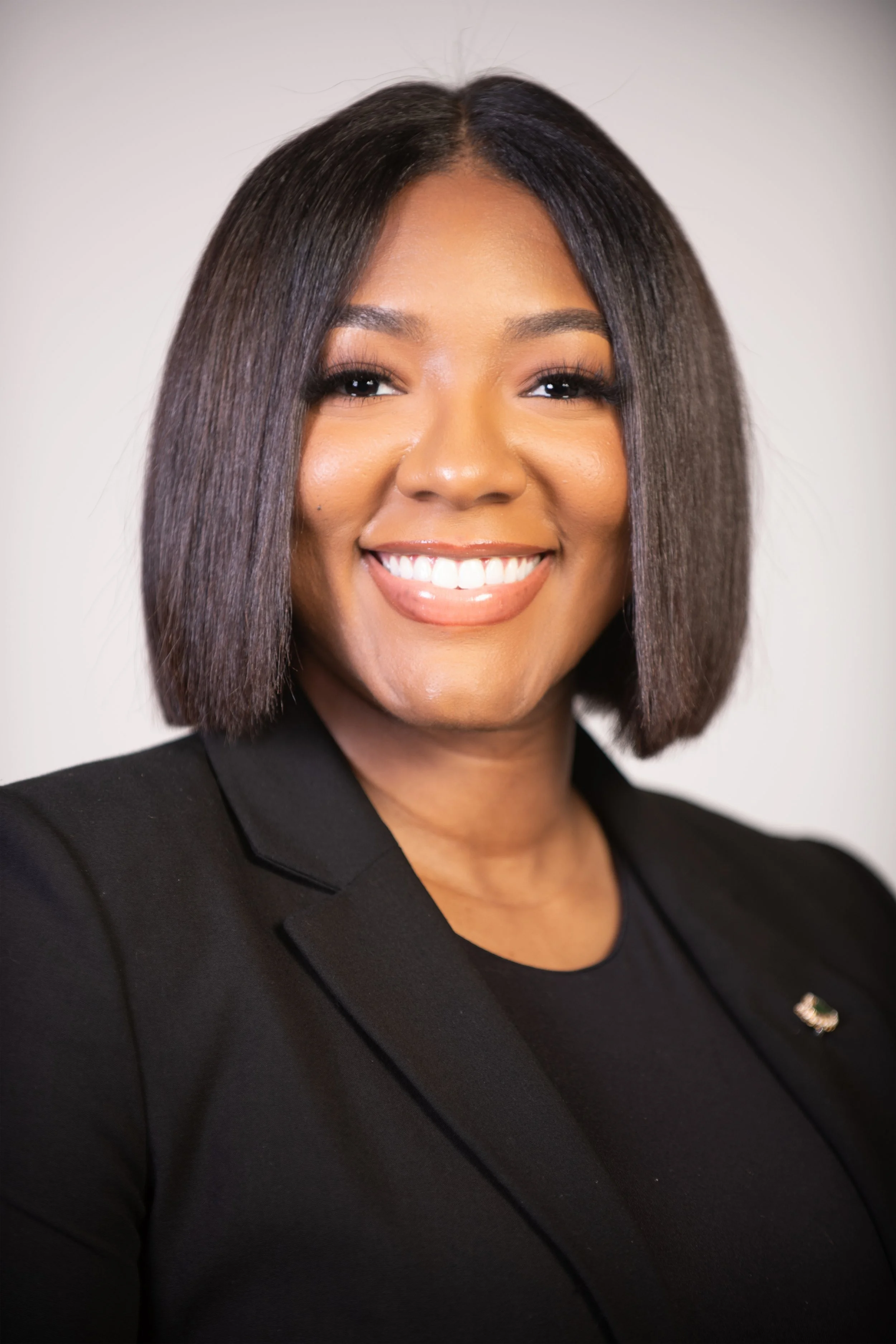 Portrait of a smiling woman with shoulder-length dark straight hair, wearing a black blazer with a pin, against a plain light background.