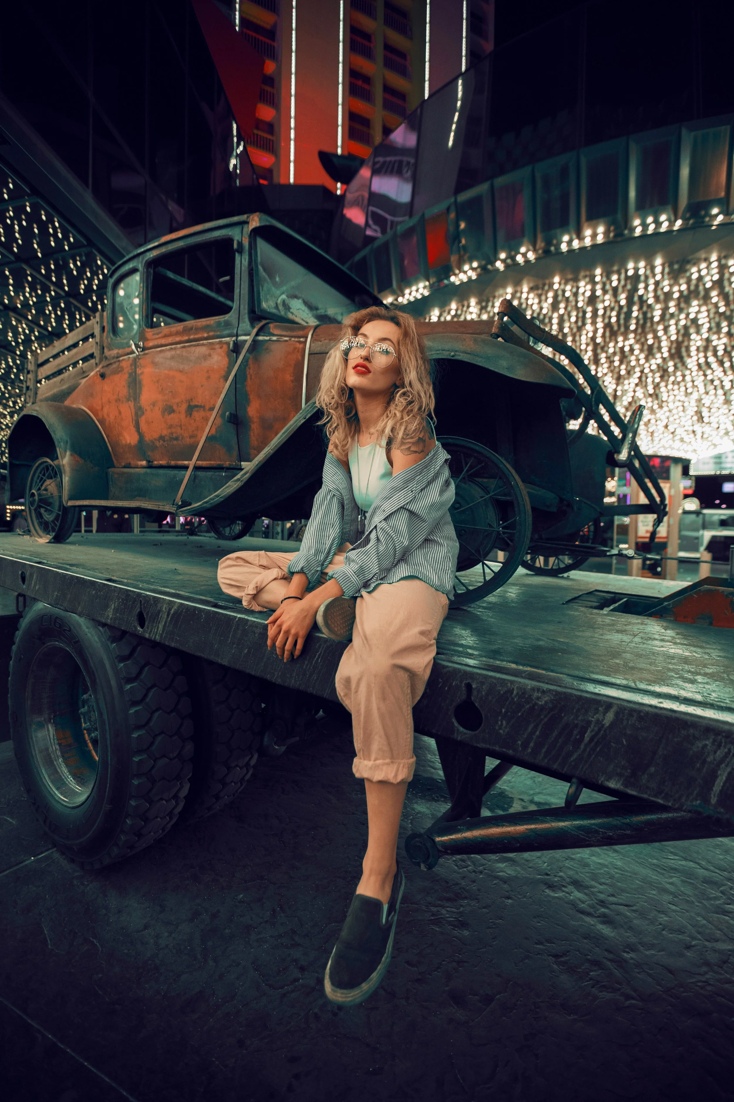 A woman with blonde curly hair, sunglasses, a white top, a blue and white striped jacket, and beige pants, sitting on the edge of a black platform in front of a rusty vintage pickup truck, illuminated by colorful city lights at night.