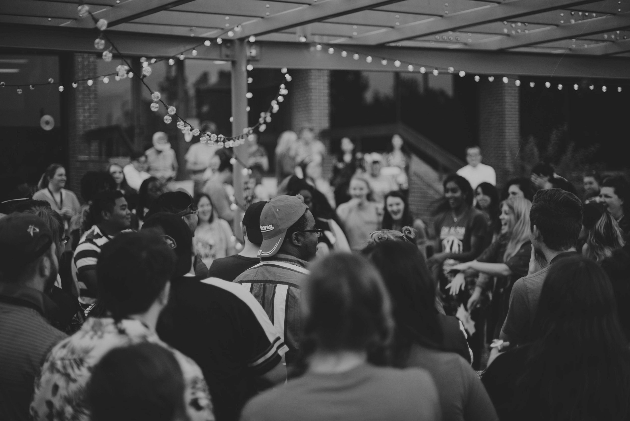Black and white photo showing a large crowd of people gathered outdoors under a covered patio with string lights, engaging in conversation and socializing.