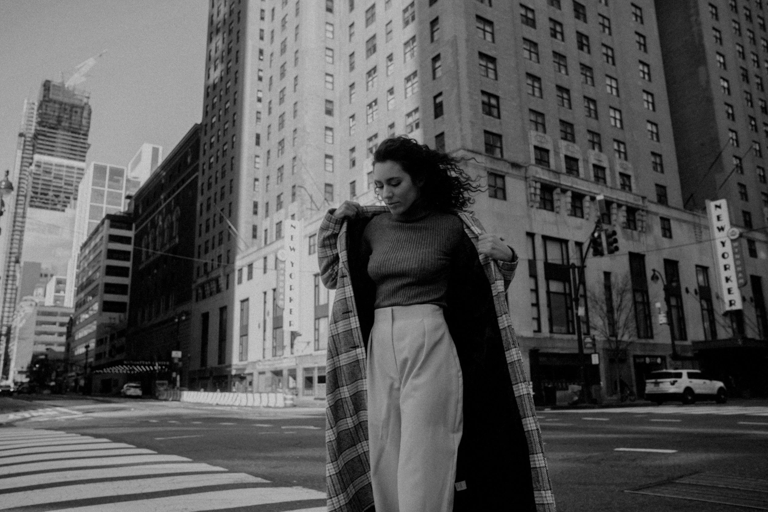 A woman walking down a city street in front of tall buildings, wearing a sweater, high-waisted pants, and a plaid coat, with her hands adjusting her coat.