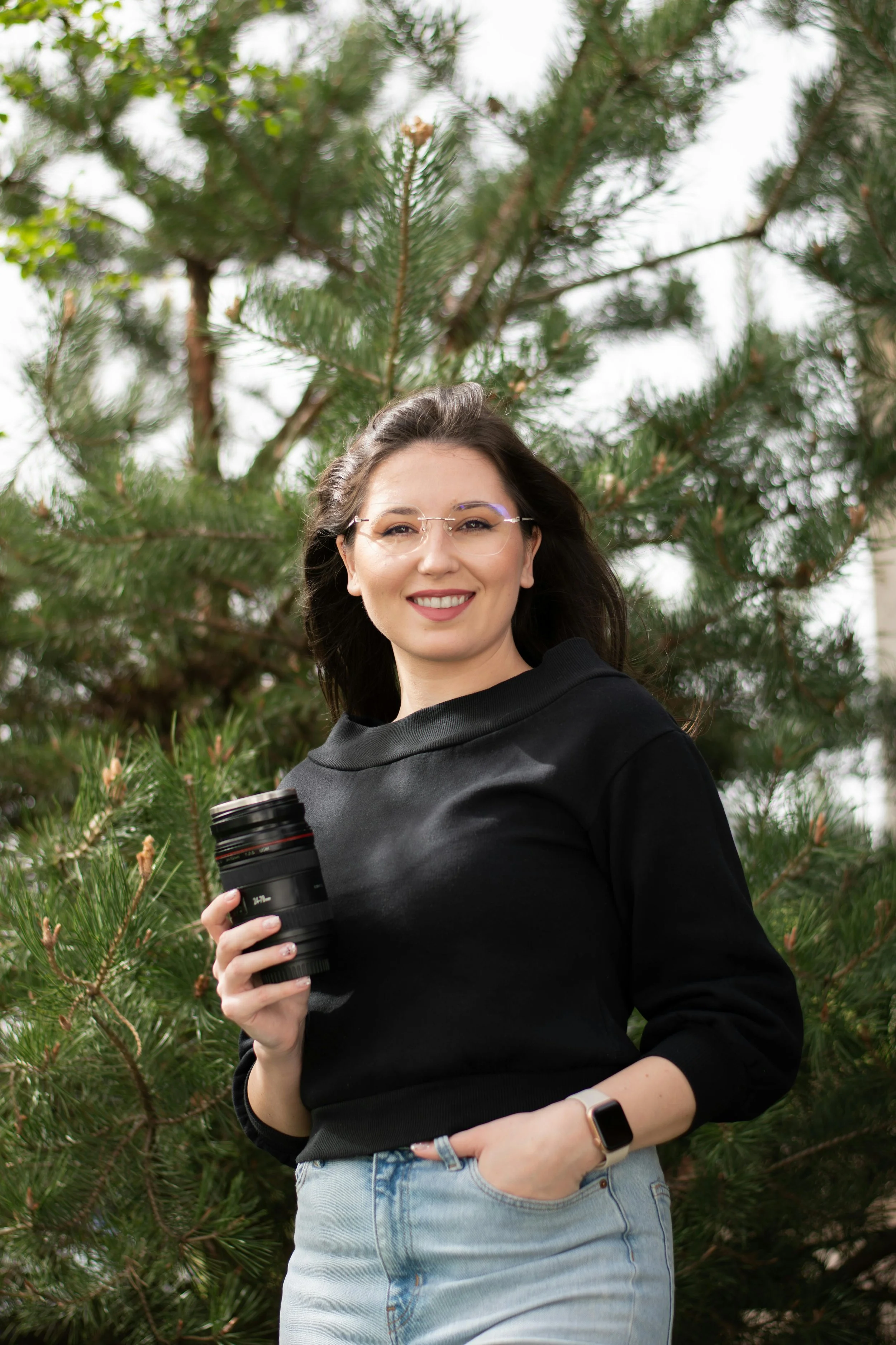 A woman with dark hair, glasses, and a black sweatshirt stands outdoors in front of a pine tree, holding a camera lens and smiling.