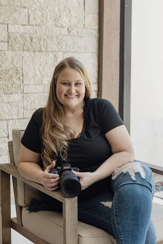 Ashley, a blond woman with long hair, sits in a beige chair holding her camera and smiling