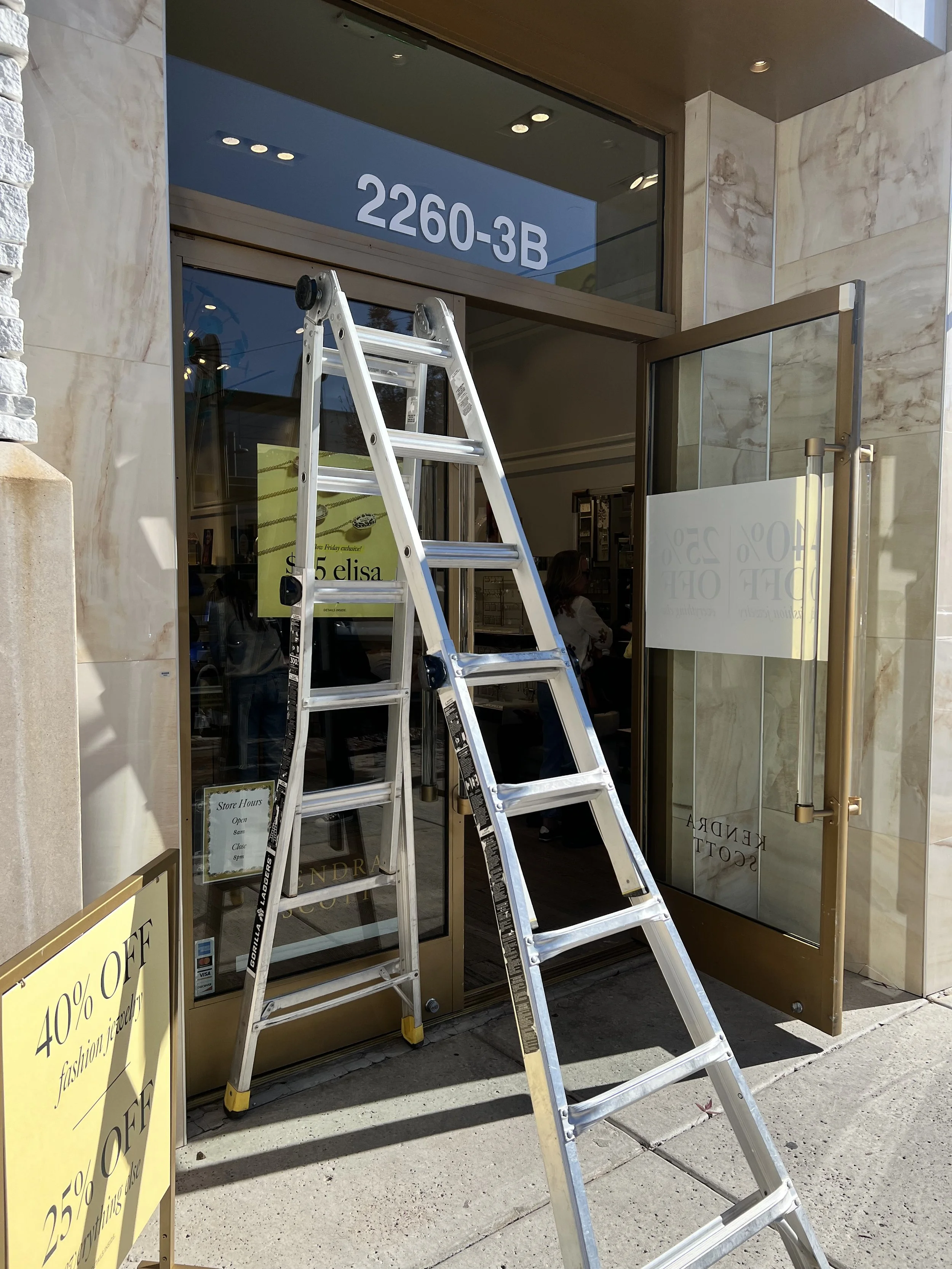 Aluminum ladder set up outside a store with glass doors, displaying store hours and promotional signs, including a yellow sign advertising 40% off.