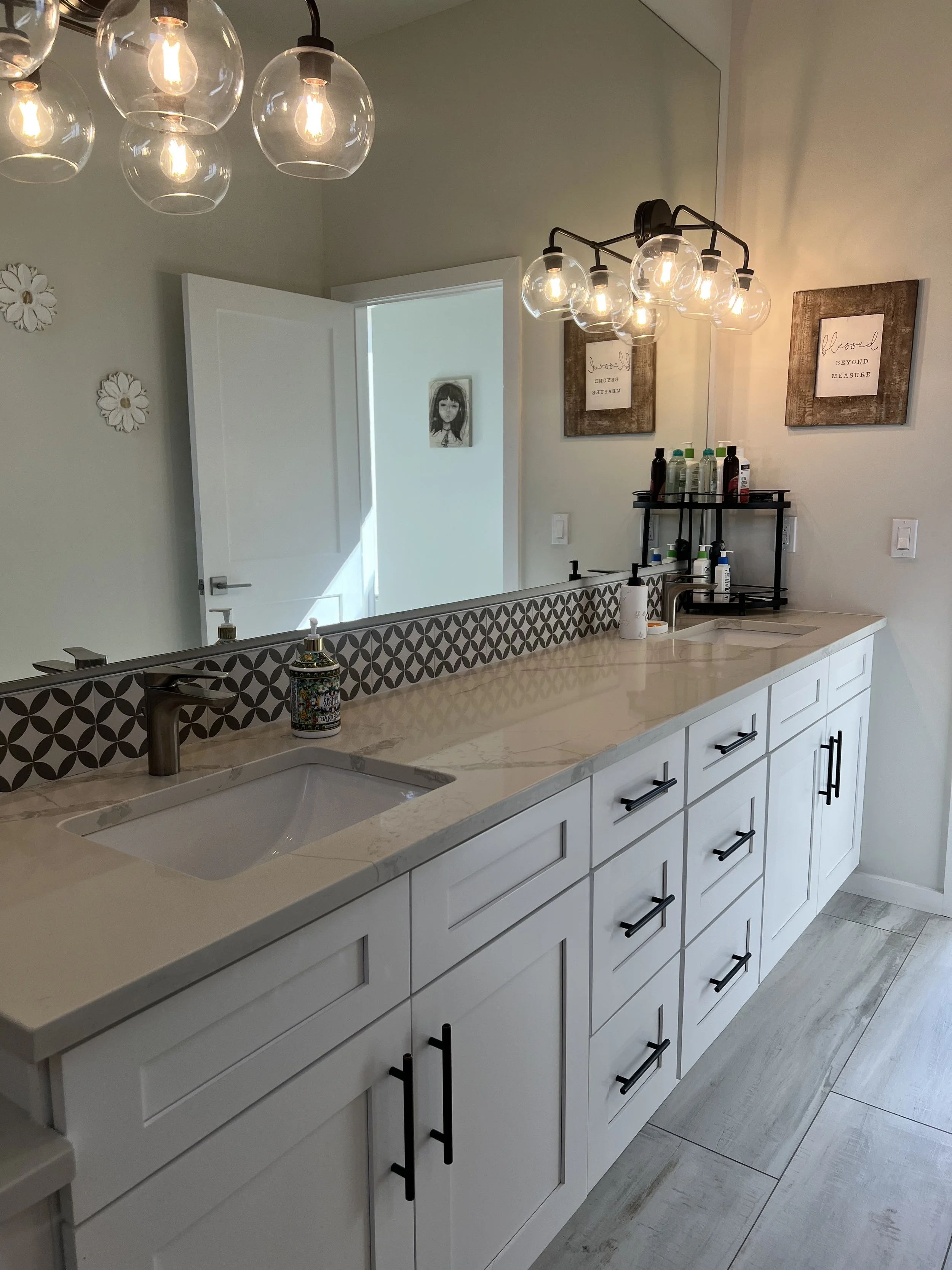 Modern bathroom vanity with white cabinetry, black handles, marble countertop, and dual sinks. Overhead lighting with multiple globe bulbs, wall decor with framed quotes, and a mirror reflecting part of the room.