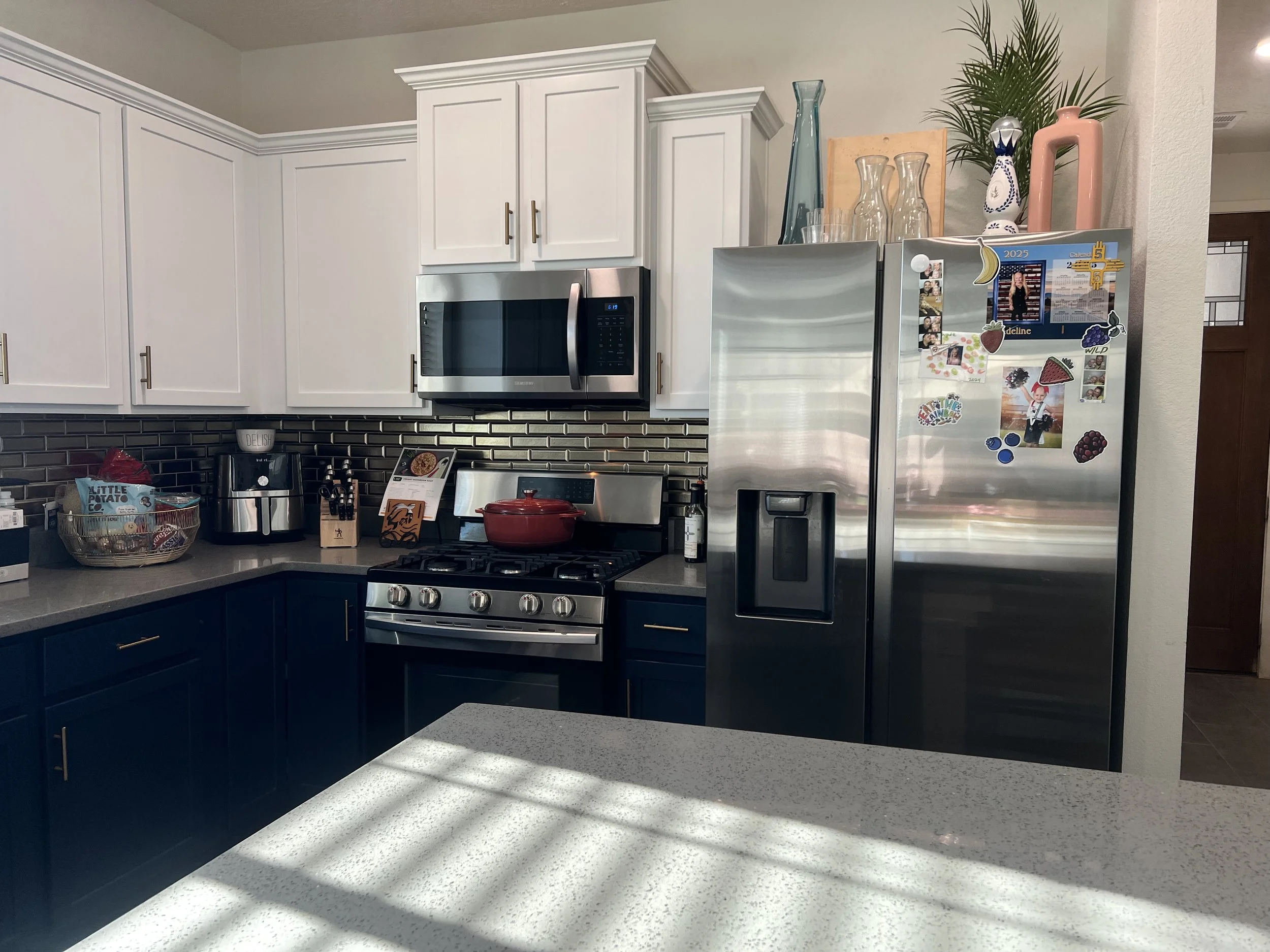 Modern kitchen with white upper cabinets, navy lower cabinets, stainless steel refrigerator, microwave, and oven. Countertop with kitchen appliances, dish rack, and decorative items.