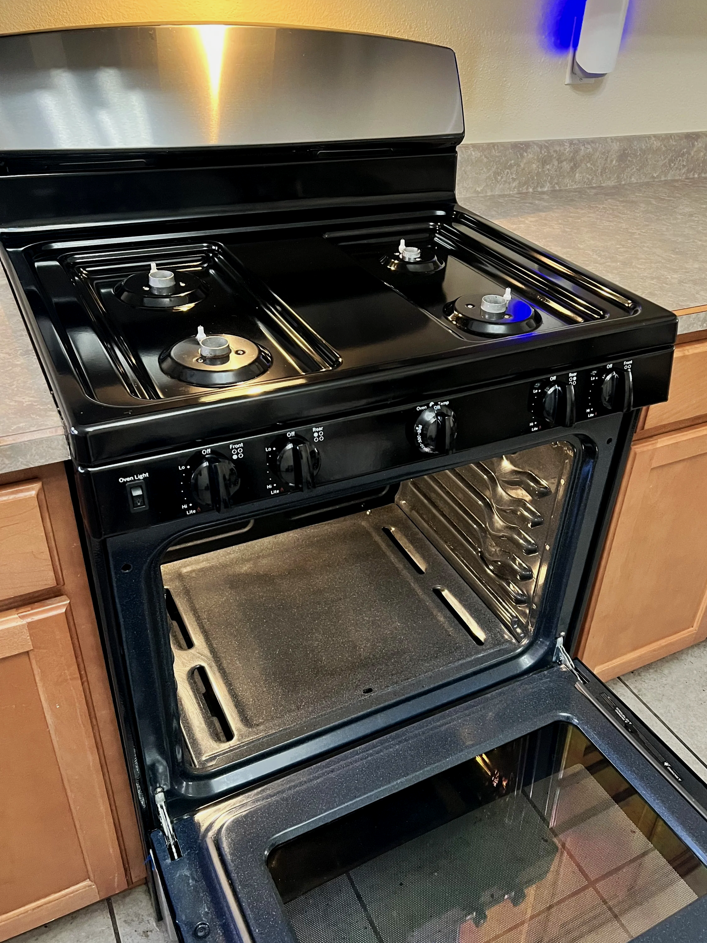Black stove with three burners and oven, with the oven door open showing the baking tray inside, in a kitchen with wooden cabinets and countertop.