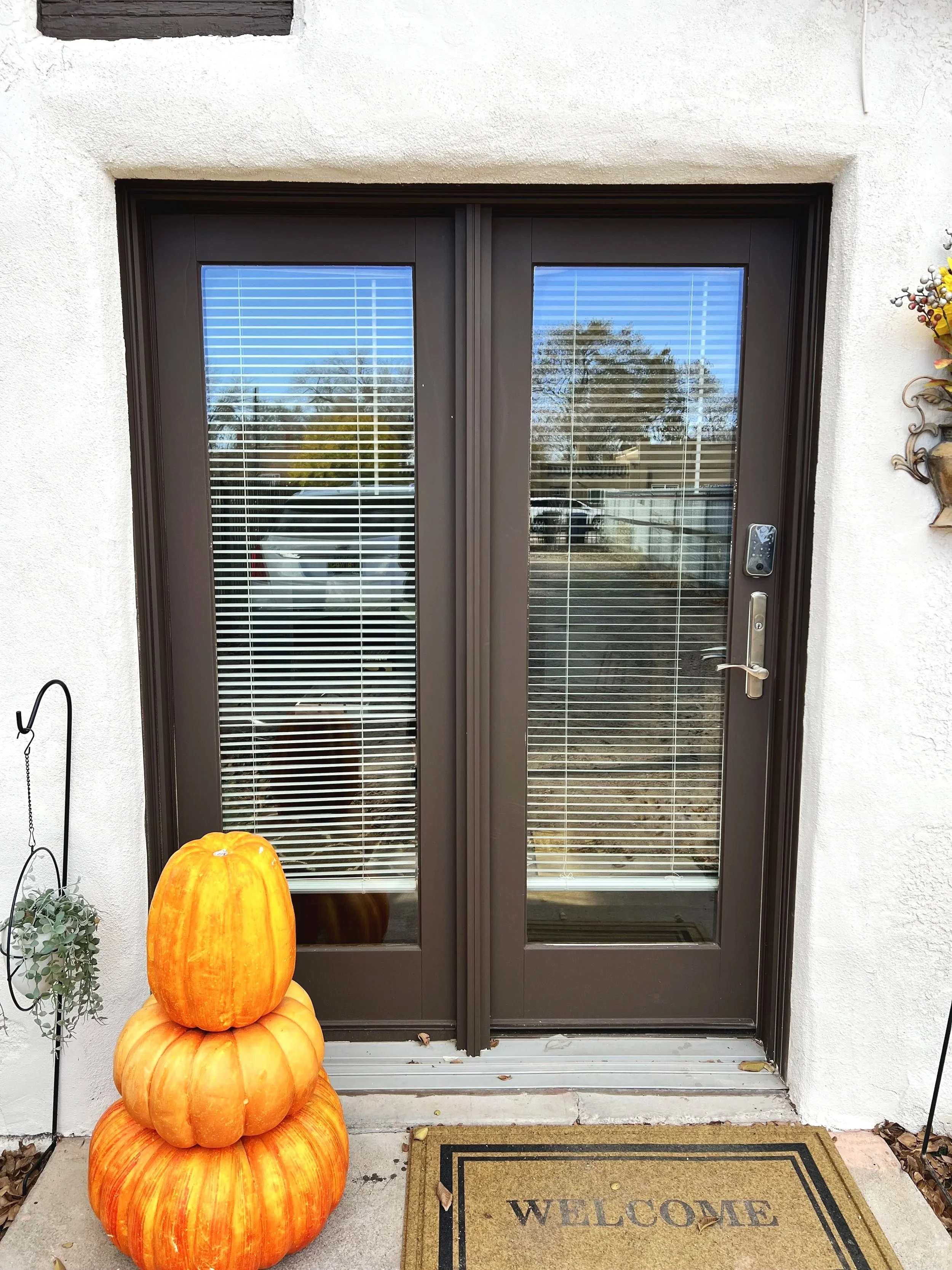 Decorative pumpkins stacked outside a front door, with a welcome mat and potted plants nearby.