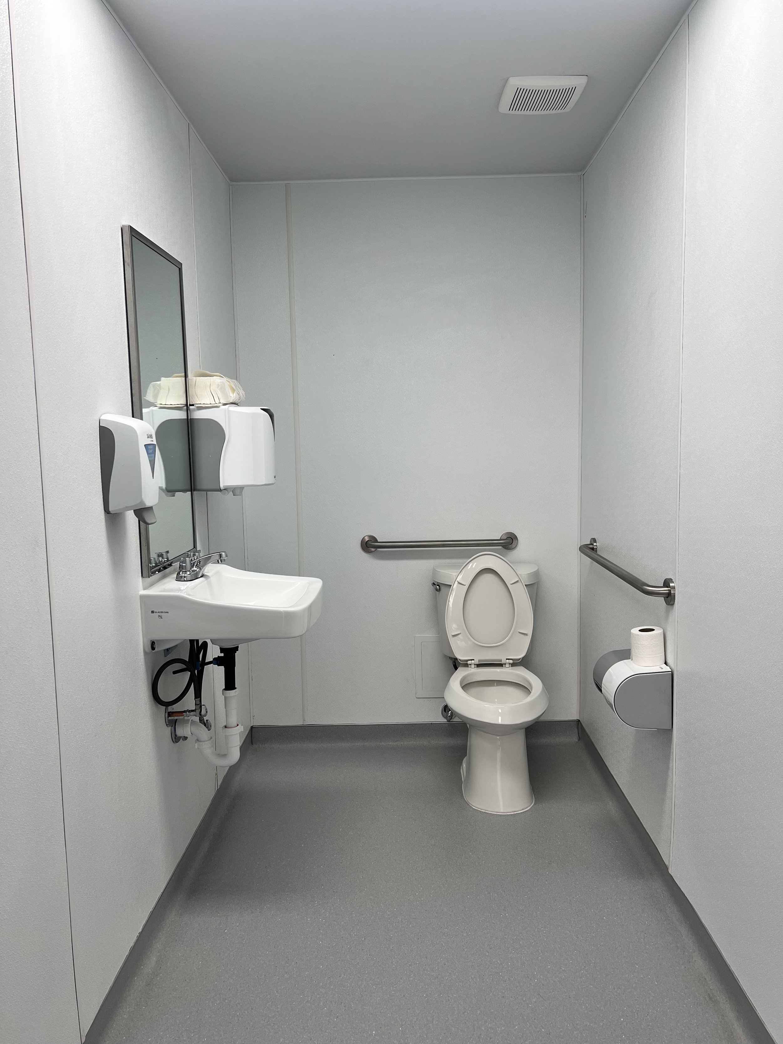 A clean, accessible public restroom with a toilet, a wall-mounted sink, a mirror, a paper towel dispenser, and handrails.