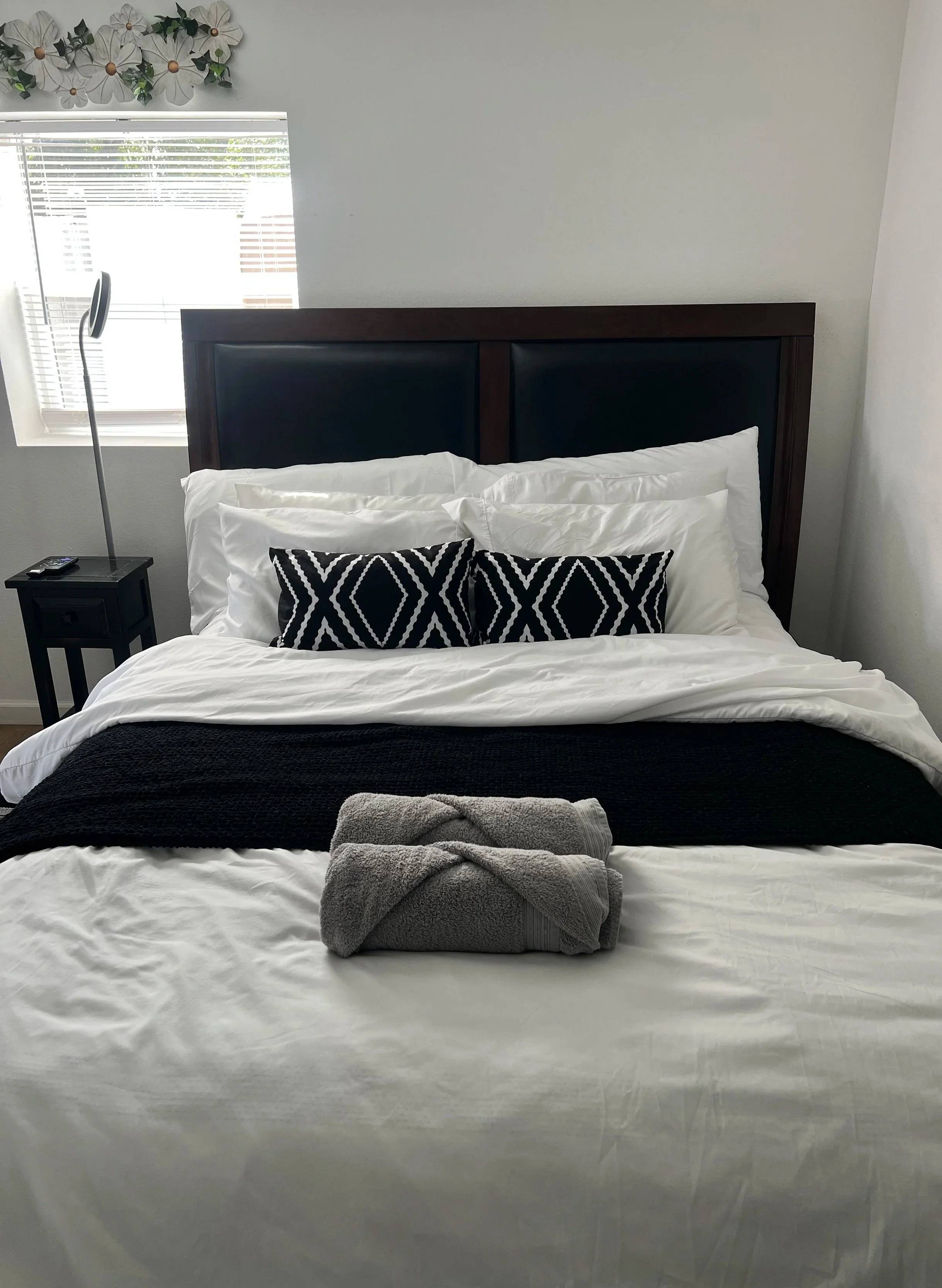 A neatly made bed with white sheets, black throw blanket, and gray towels on top, in front of a headboard with black cushions. There are two patterned decorative pillows, and a nightstand with a remote control and a modern silver lamp. A window with blinds and a floral wall decoration are in the background.