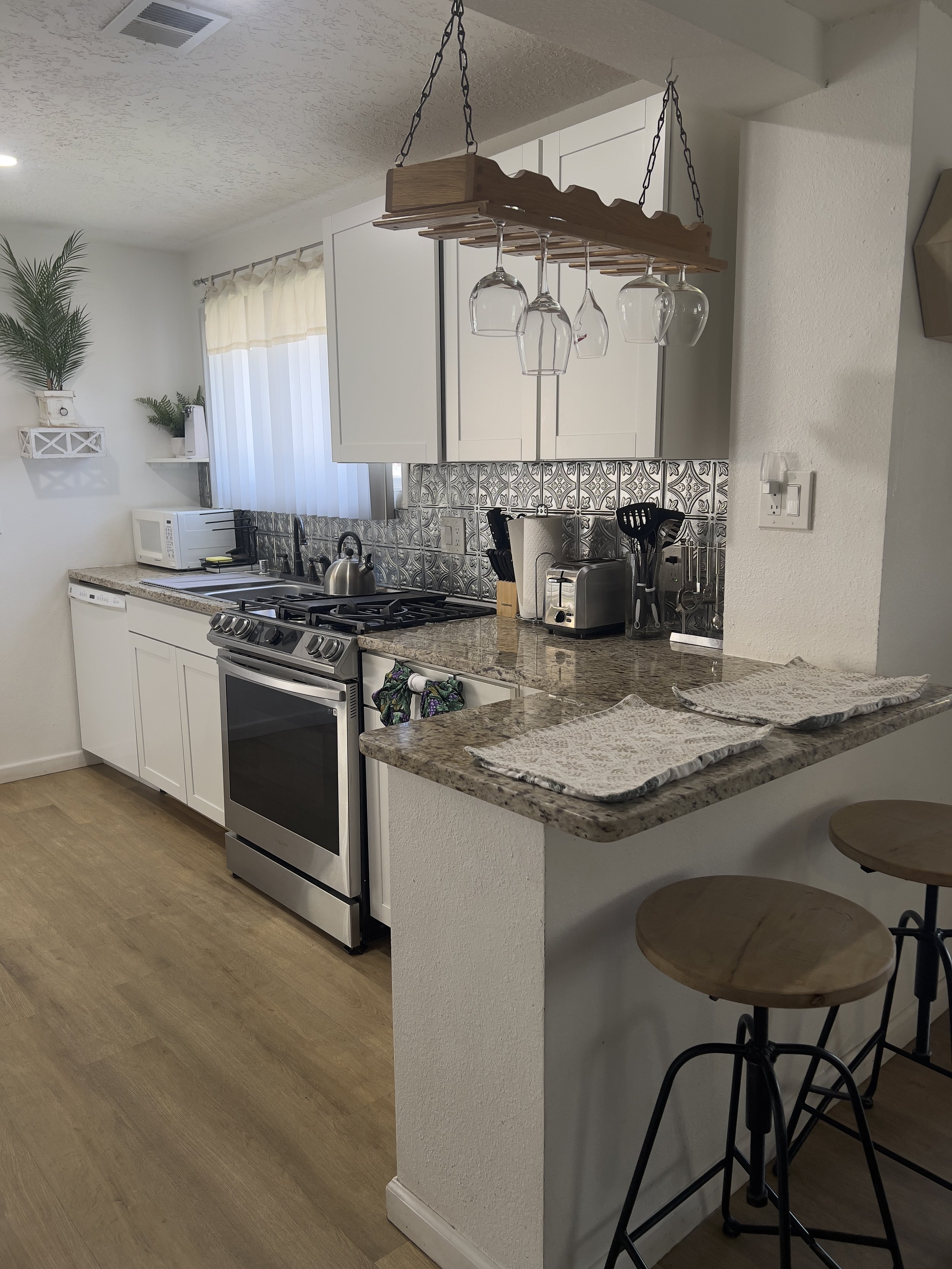 Kitchen with white cabinets, granite countertops, and a patterned tile backsplash. Includes a stove, microwave, toaster, and kitchen utensils. Over-the-counter wine glass rack, two bar stools at a breakfast bar, and decorative plants with a window with cream-colored curtains.