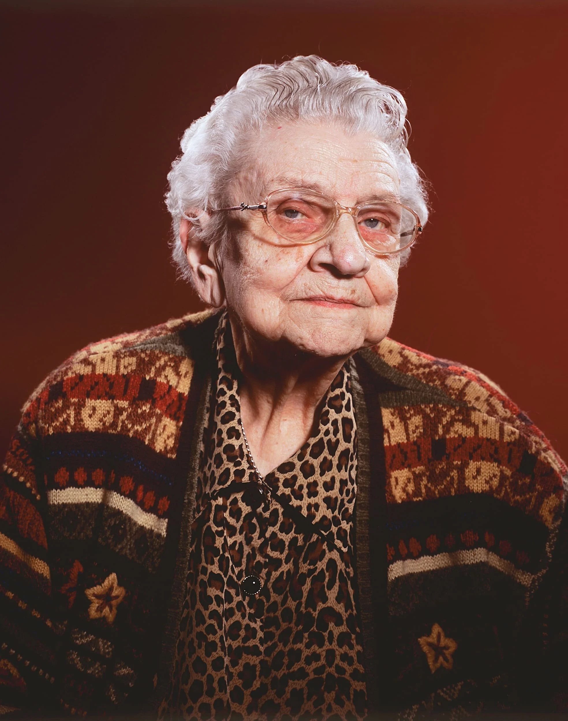 An elderly woman with white hair and glasses, wearing a patterned sweater and a leopard print shirt, looking slightly to the side against a brown background.