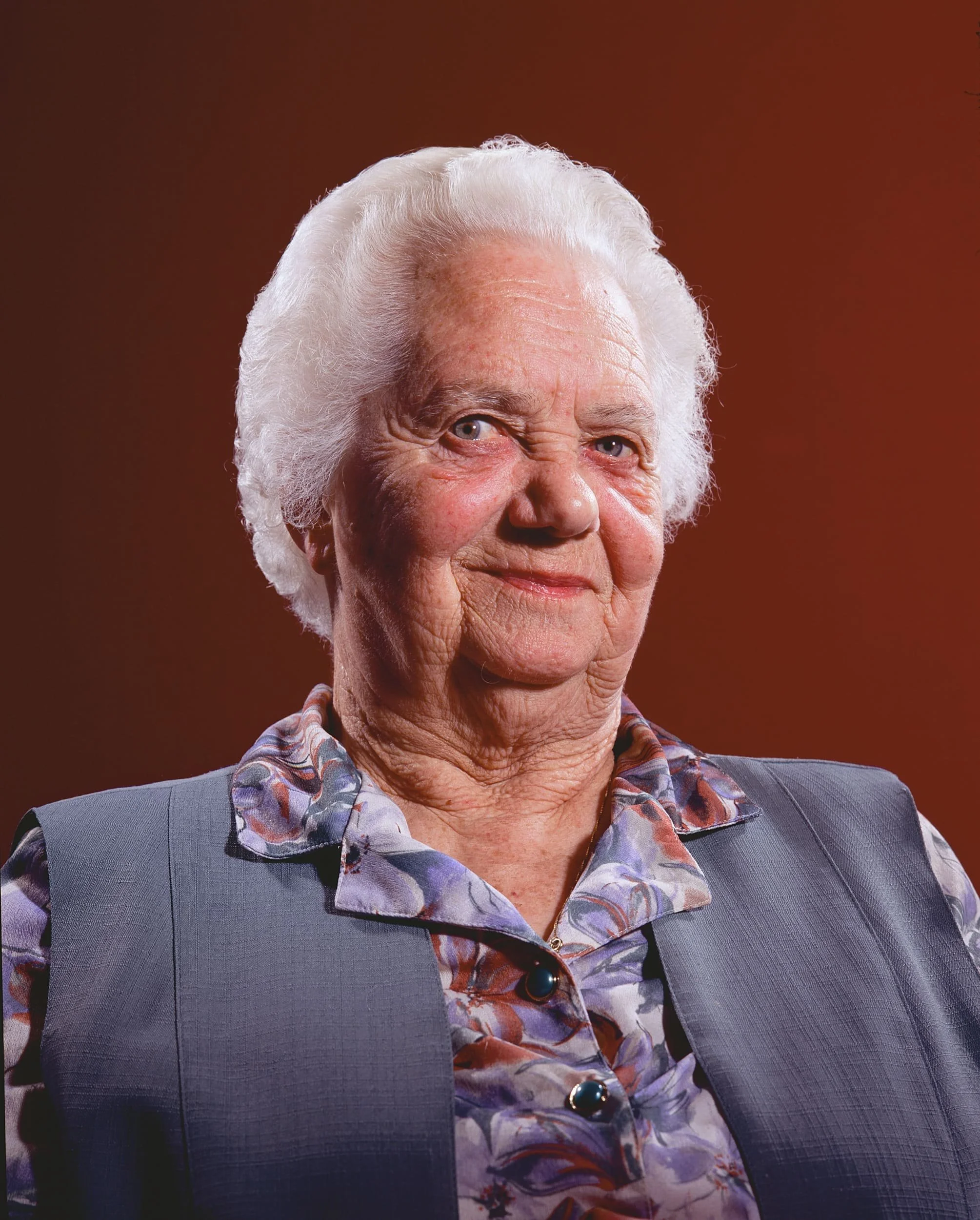 Portrait of an elderly woman with white hair, wearing a floral blouse and a gray vest, smiling slightly against a dark red background.