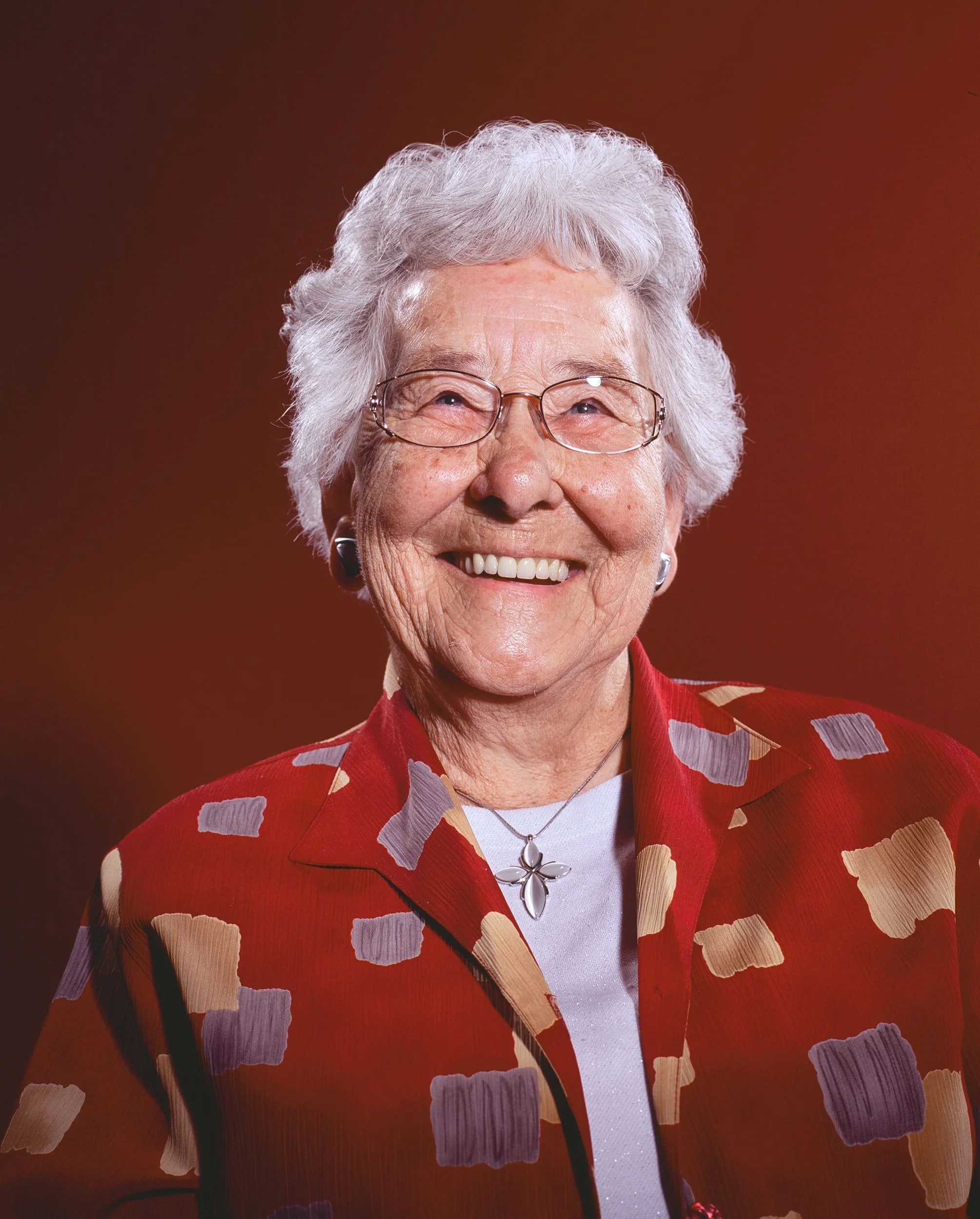 An elderly woman with gray hair styled in soft curls, smiling and wearing glasses. She has on a red patterned blazer with purple, beige, and gray shapes, a white top underneath, a silver necklace with a floral pendant, and earrings. The background is