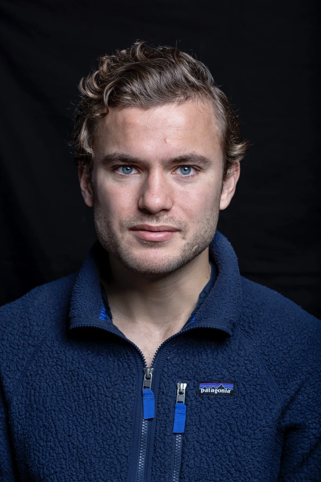 A young man with short curly blond hair, blue eyes, and light facial hair wearing a navy blue Patagonia fleece jacket against a black background.