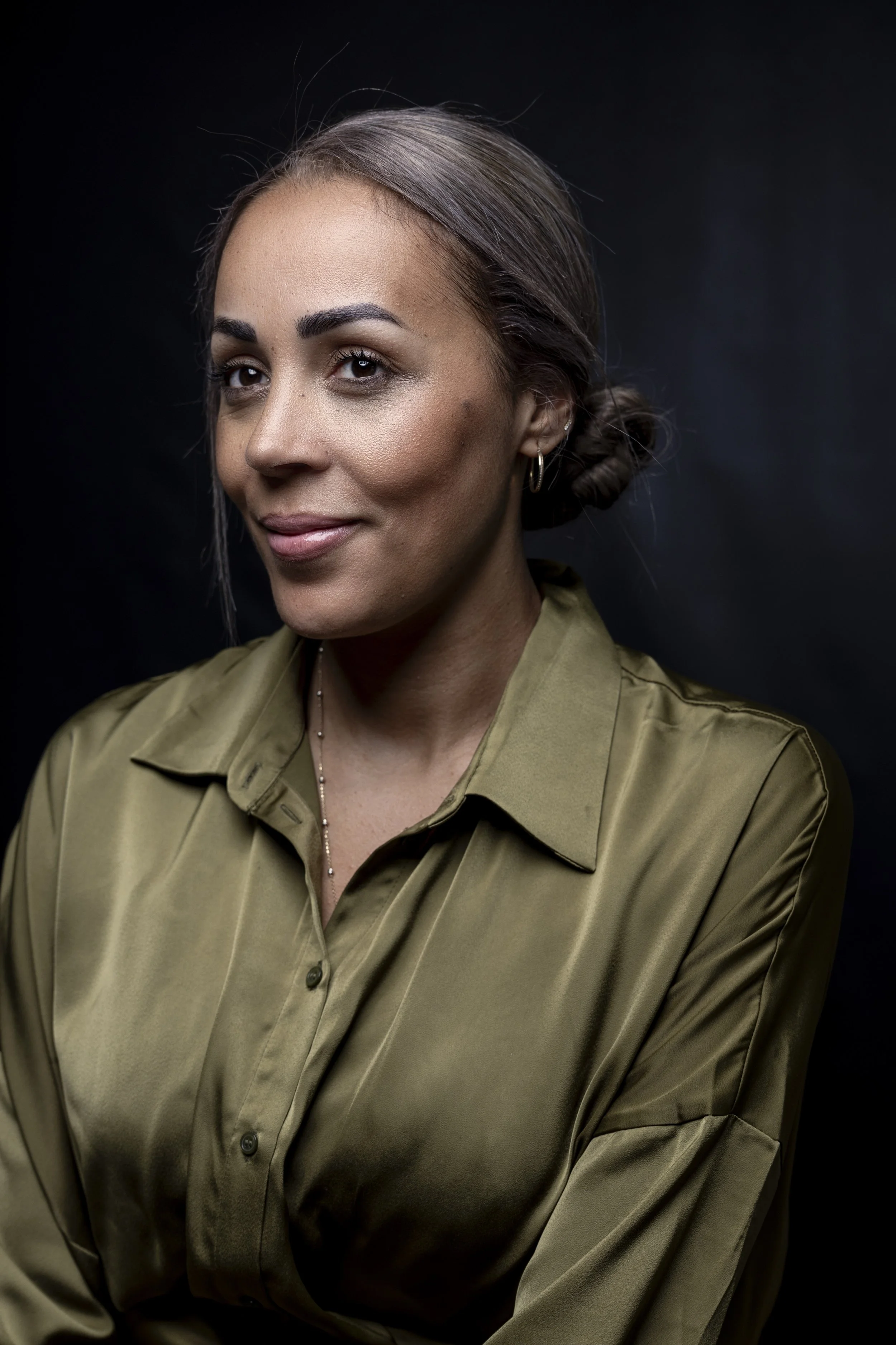 Portrait of a woman with light brown hair styled in a bun, wearing a green satin blouse with buttons, small hoop earrings, and a delicate necklace, against a black background.