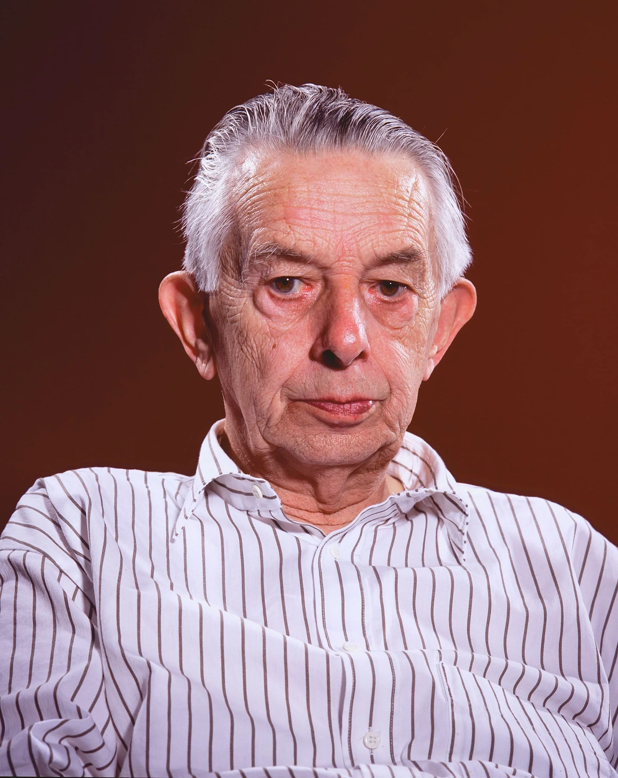 Close-up of an elderly man with gray hair, wearing a white and brown striped button-up shirt, against a brown background.