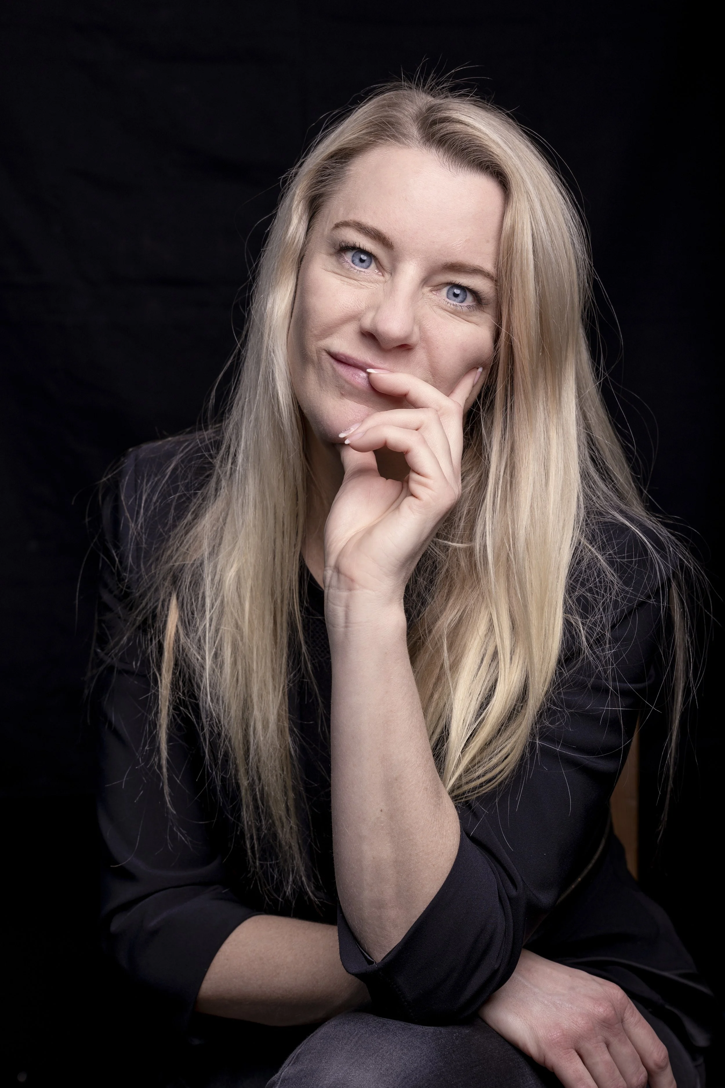 A woman with long blonde hair and blue eyes, wearing a black top, gazes softly at the camera with her hand resting on her chin, against a dark background.