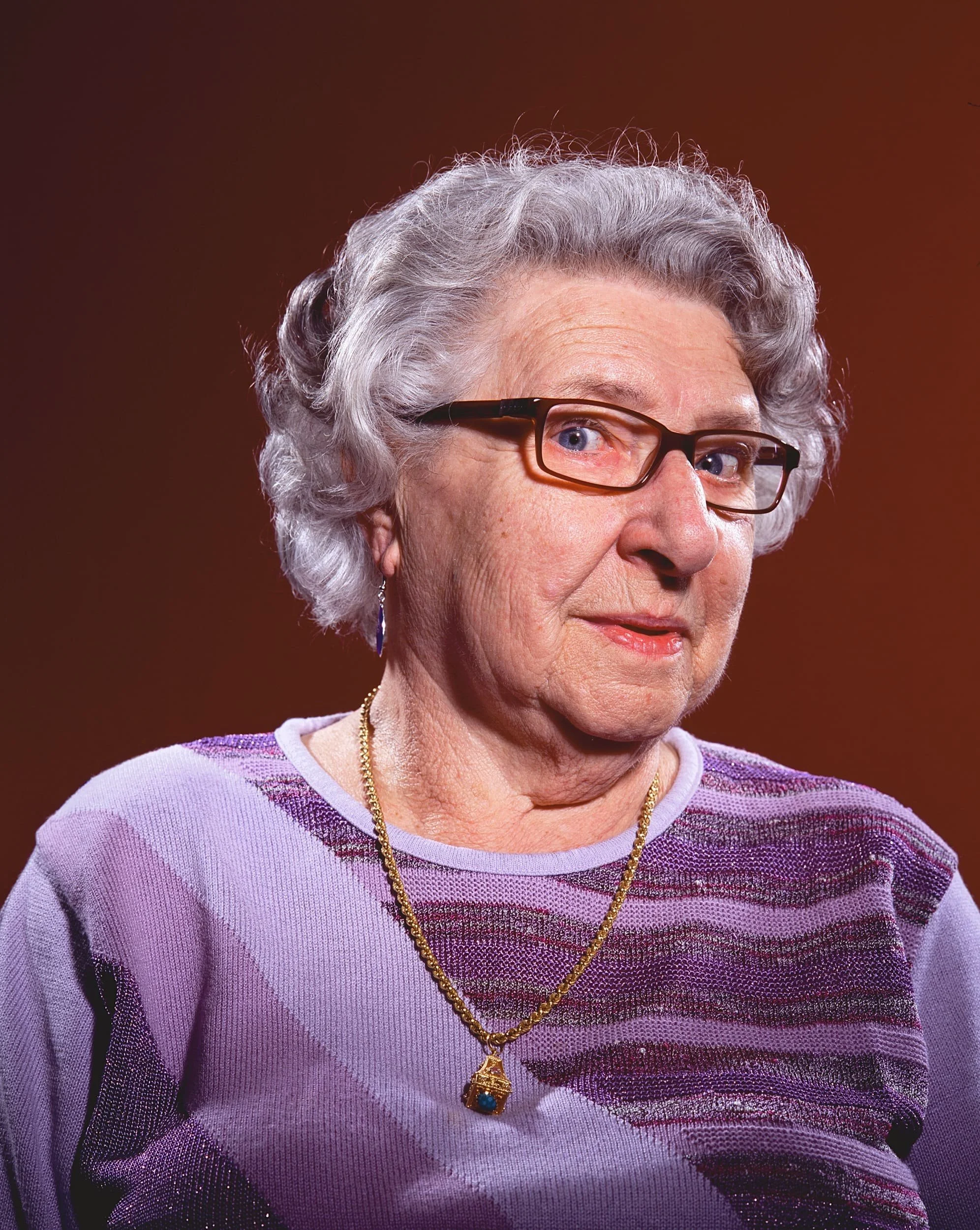 A portrait of an elderly woman with gray hair, wearing glasses, a purple and gray striped top, and a gold necklace with a blue pendant, against a brown background.