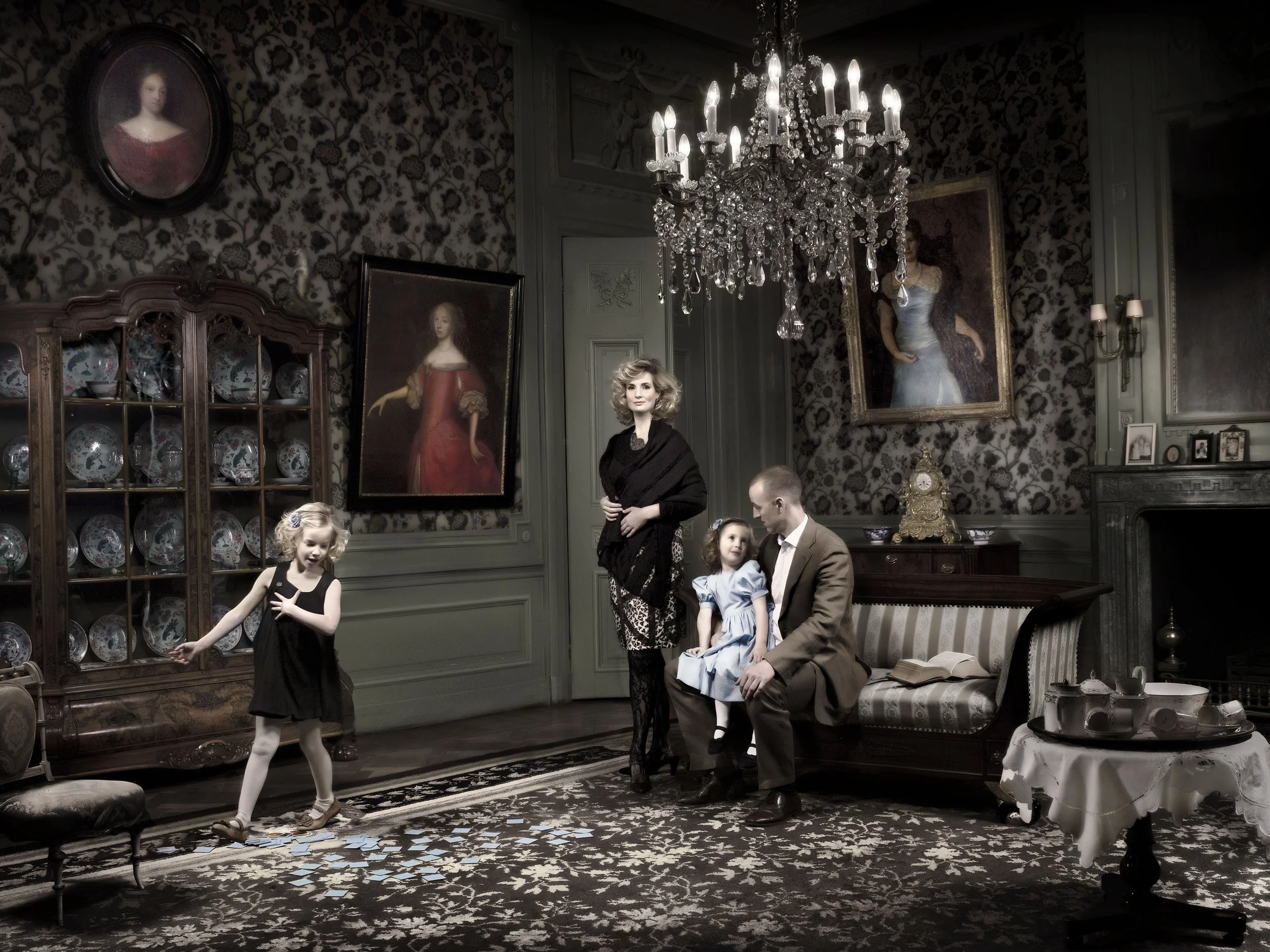 A vintage-styled living room with a chandelier, wallpaper, and portraits, featuring a woman, three children, and a man sitting on a couch, two children playing with cards or paper on the floor.