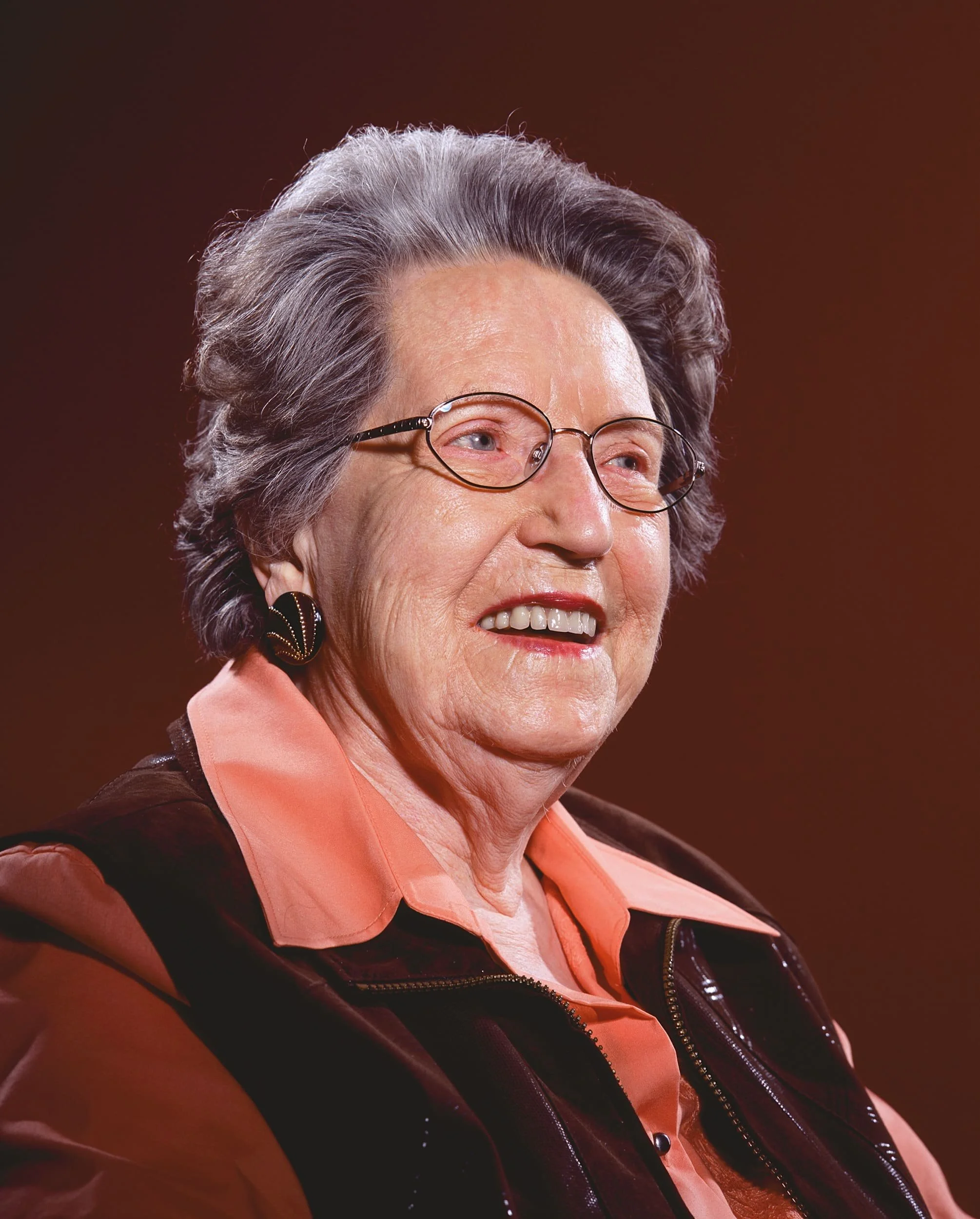 An elderly woman with gray hair and glasses, smiling, wearing a peach-colored shirt and a dark vest.