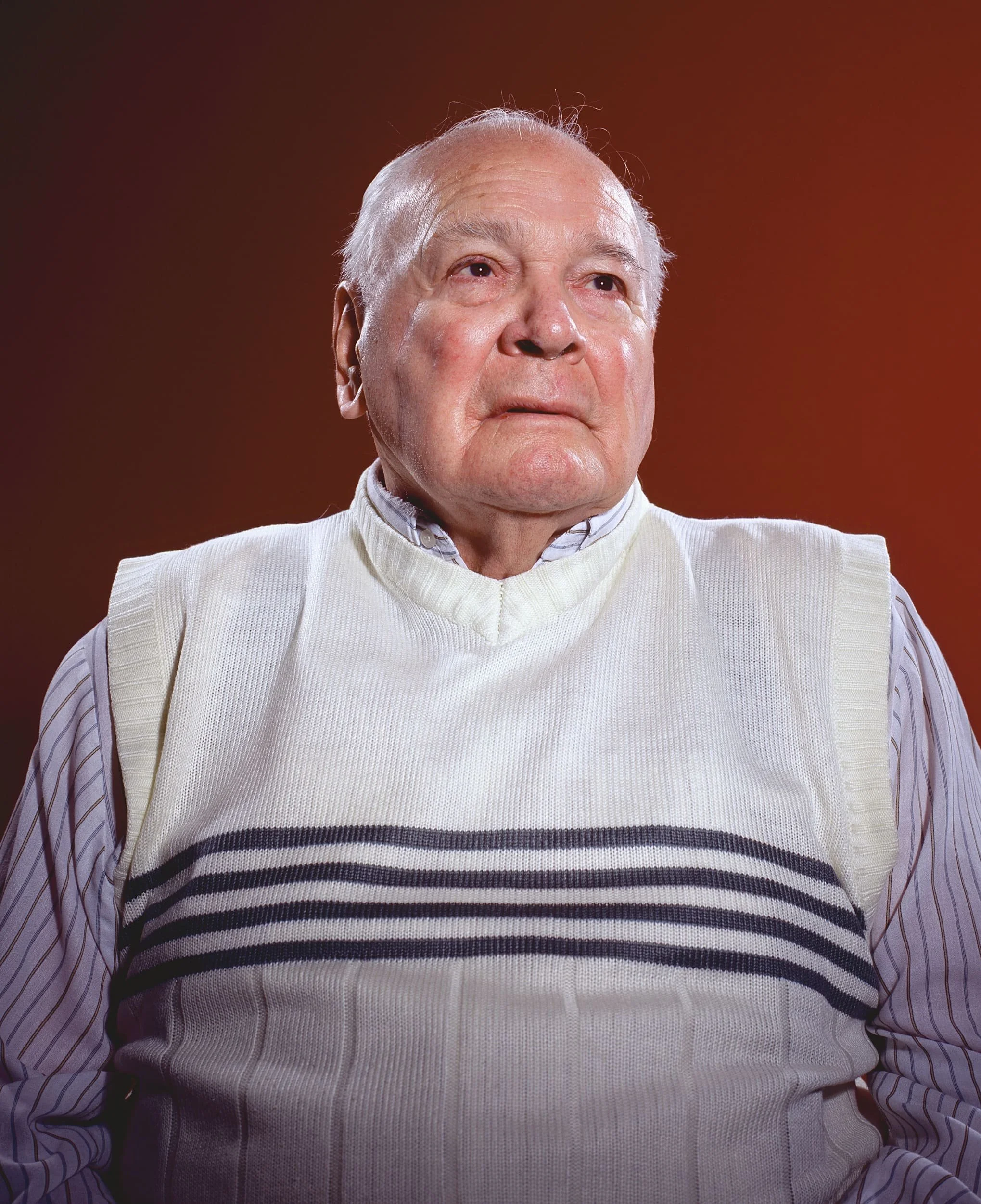 Portrait of an elderly man with a serious expression, wearing a white vest with black stripes over a striped shirt, against a warm reddish background.