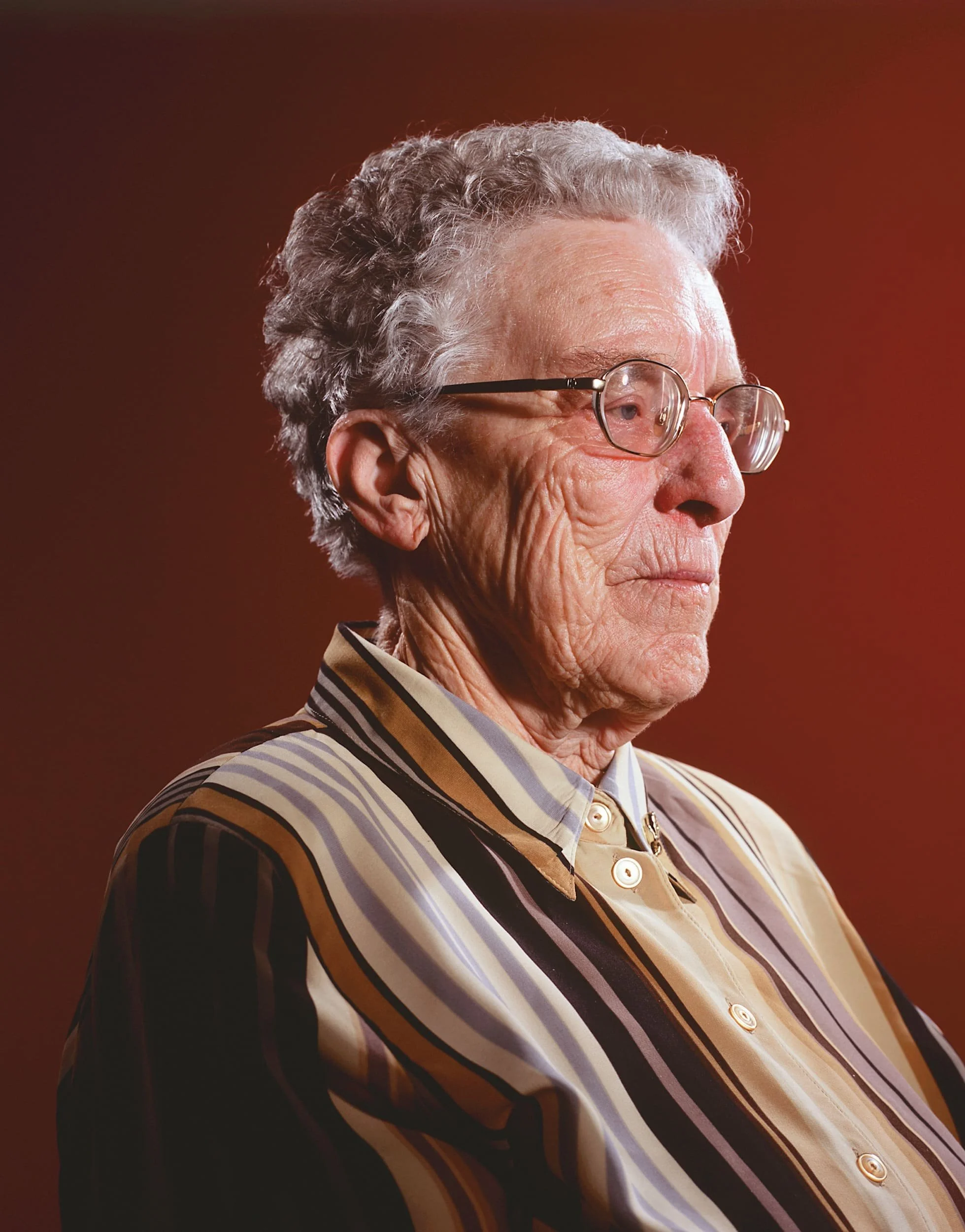 Close-up of an elderly woman with gray hair, wearing glasses, and a multicolored striped shirt, looking to the side against a warm brown background.