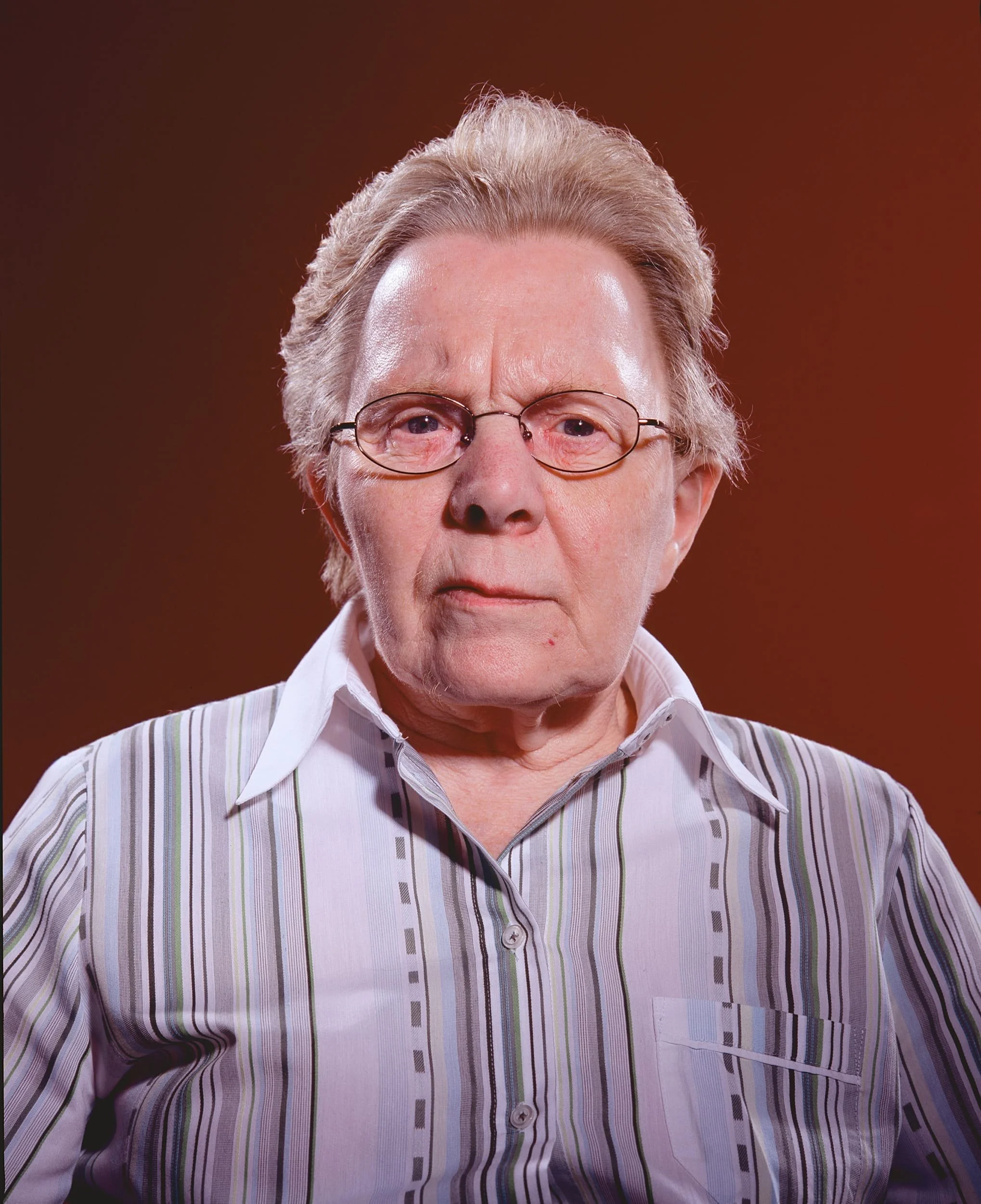 Close-up of an older man with gray hair, glasses, wearing a striped shirt, looking serious against a brown background.