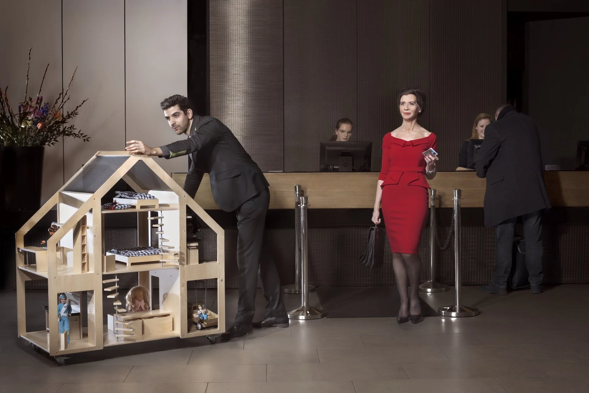 Two hotel staff members organizing a dollhouse in the lobby of a hotel, with a woman in a red dress waiting at the check-in counter in the background.