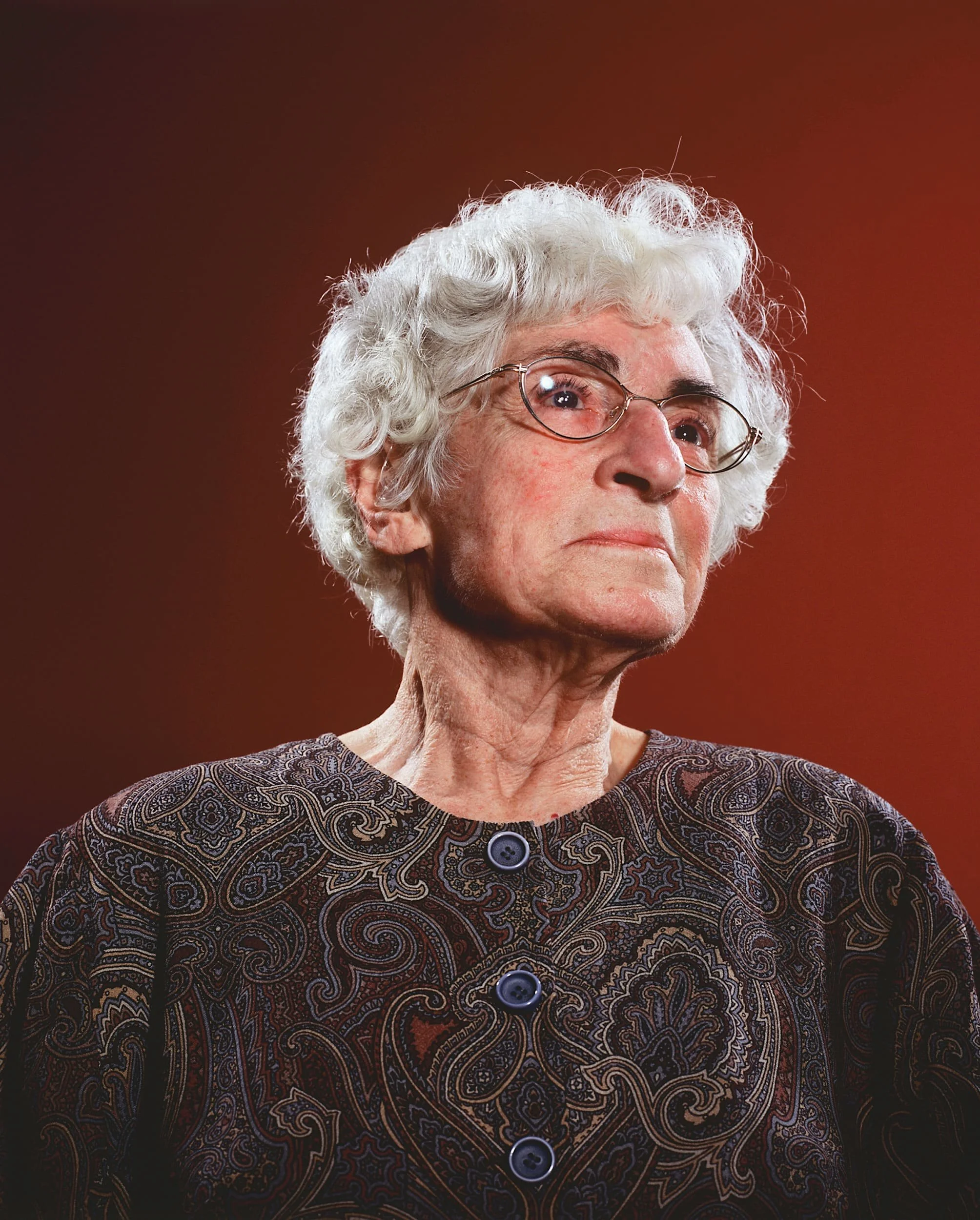 An elderly woman with white curly hair and glasses, wearing a dark patterned top, looking to her left against a reddish-brown background.