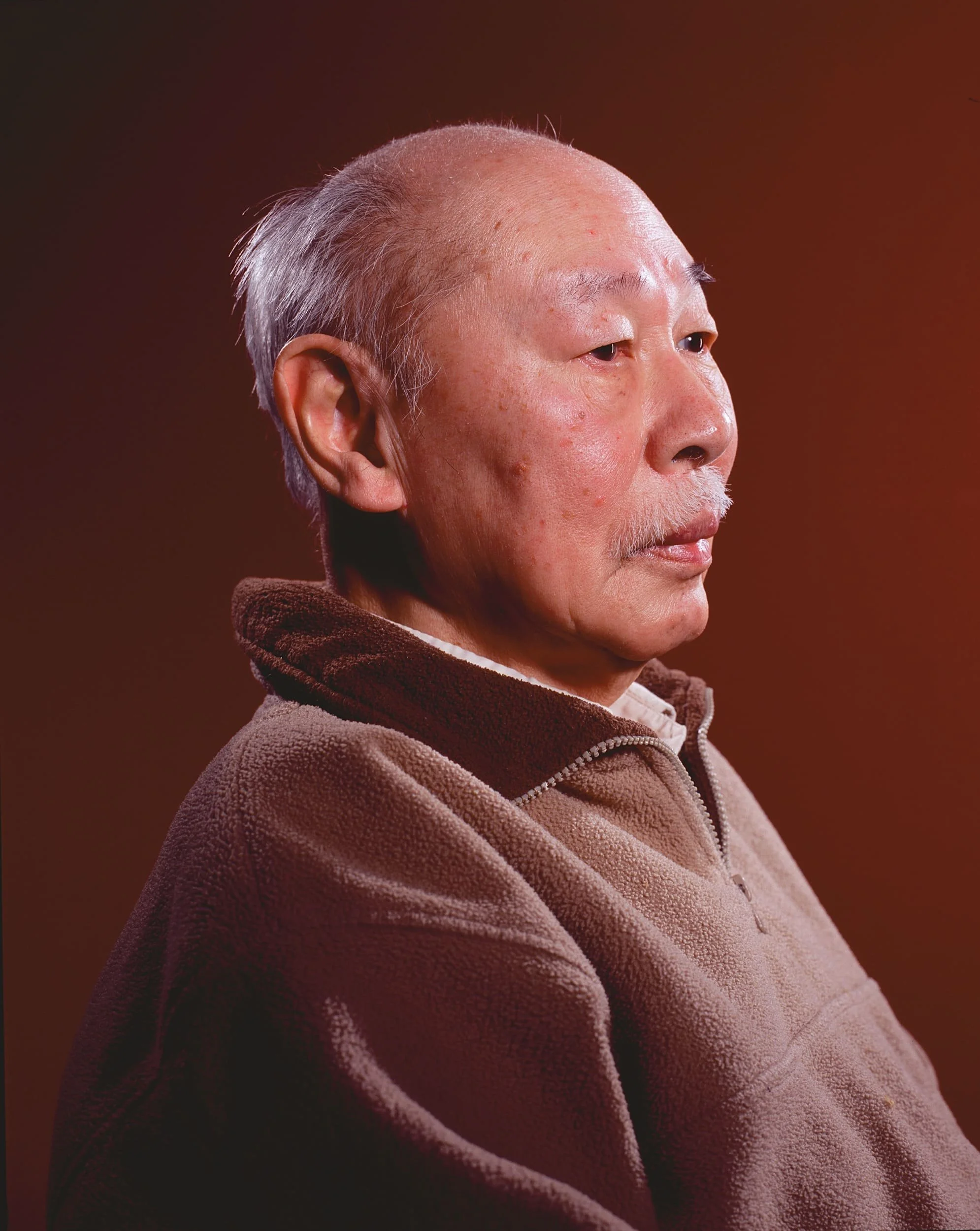 Profile of an elderly Asian man with gray hair and mustache, wearing a brown fleece jacket, against a dark red background.