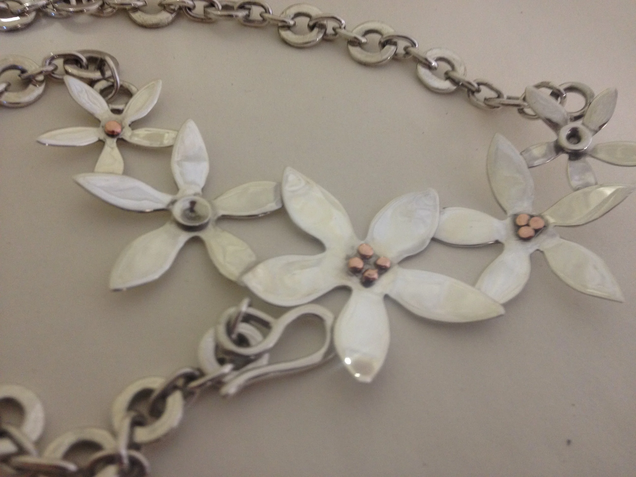 A silver chain necklace with flower-shaped pendants made of white shell-like material, each with a small pink bead in the center, on a light gray surface.
