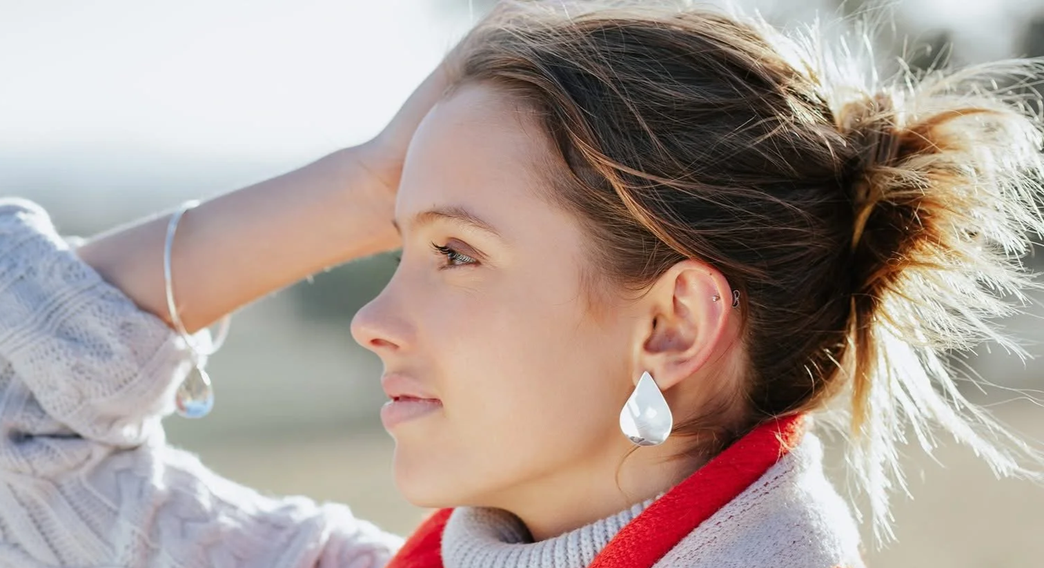 Profile of a young woman with her hand on her head, wearing large teardrop earrings and a white sweater with a red hood, outdoors in daylight.