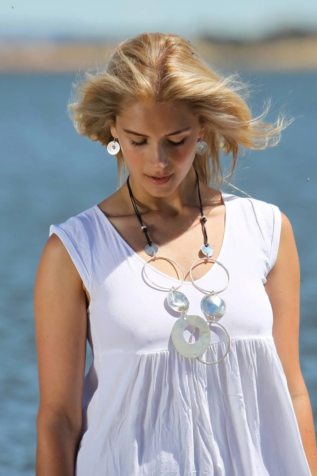 A woman with blonde hair stands outdoors near water, wearing a white dress and large silver jewelry, including earrings and a statement necklace.