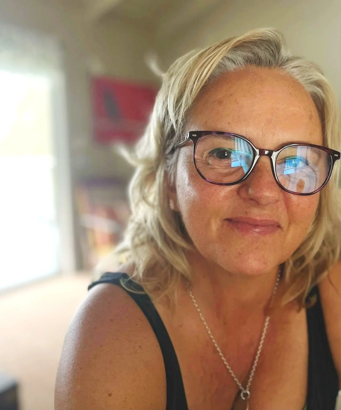 A woman with blonde curly hair, wearing glasses and a black sleeveless top, smiles in a close-up selfie. She is wearing a silver necklace with a pendant.