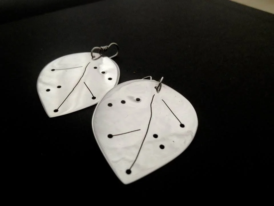 Pair of white shell earrings with black dot and line designs, placed on a dark surface.