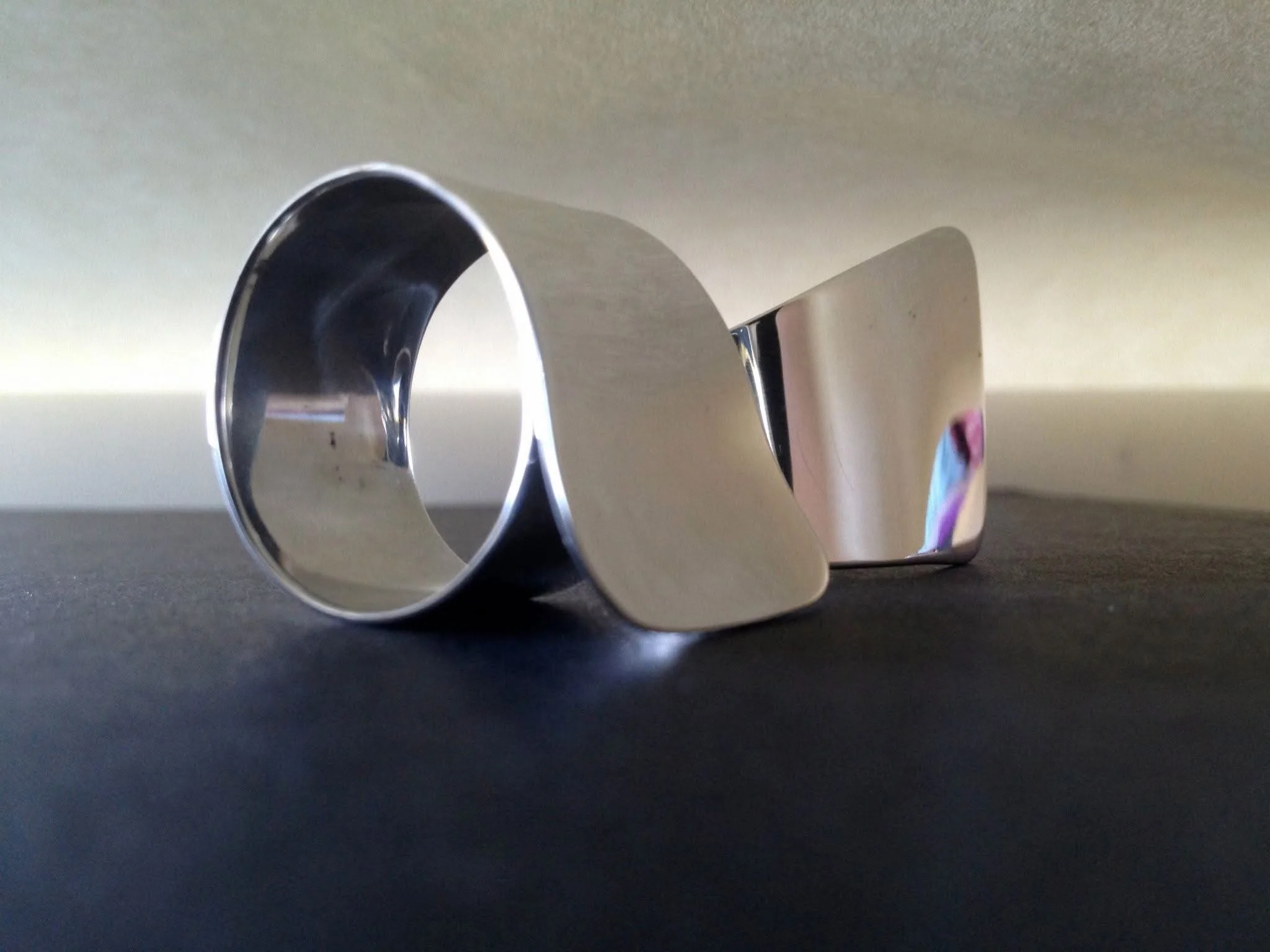 Close-up of a metallic object with a curved shape, possibly a reflective tube or handle, placed on a dark surface with a plain light-colored wall in the background.