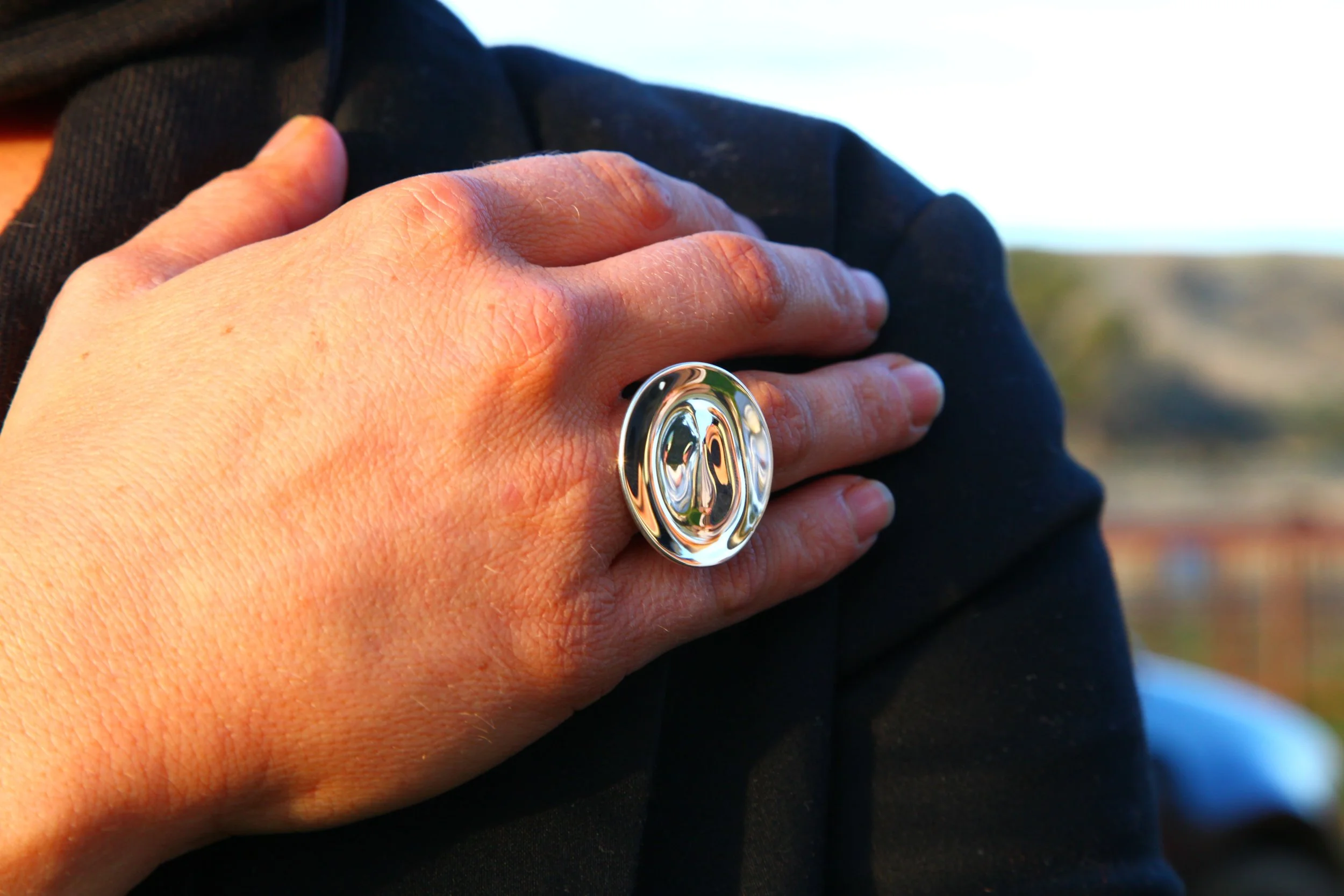 Close-up of a person's hand with a shiny silver ring, resting on their chest, wearing a black garment, with an outdoor background.