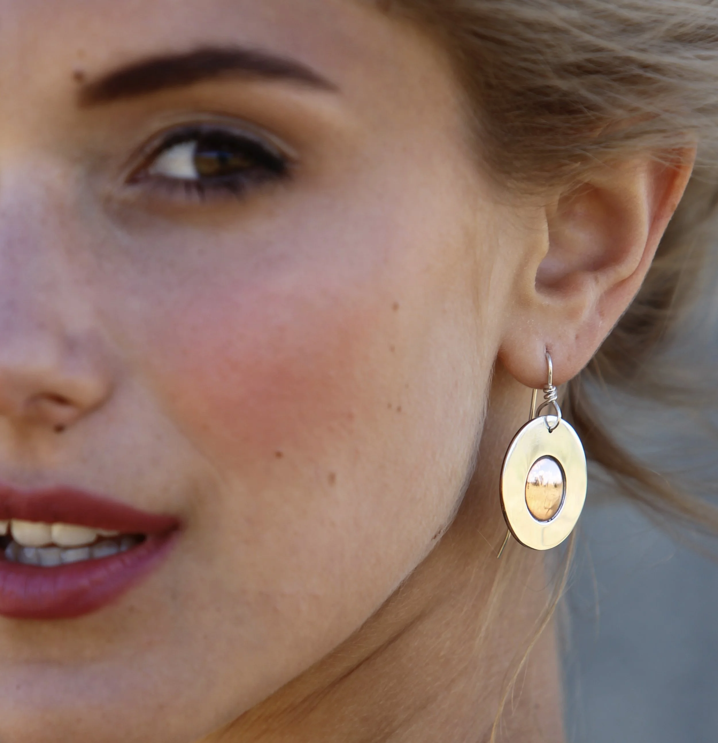 Close-up of a woman's eye, showing her eyebrow, eyelid, and part of her cheek; she has fair skin and is wearing a large, round, metallic earring with a reflective surface.
